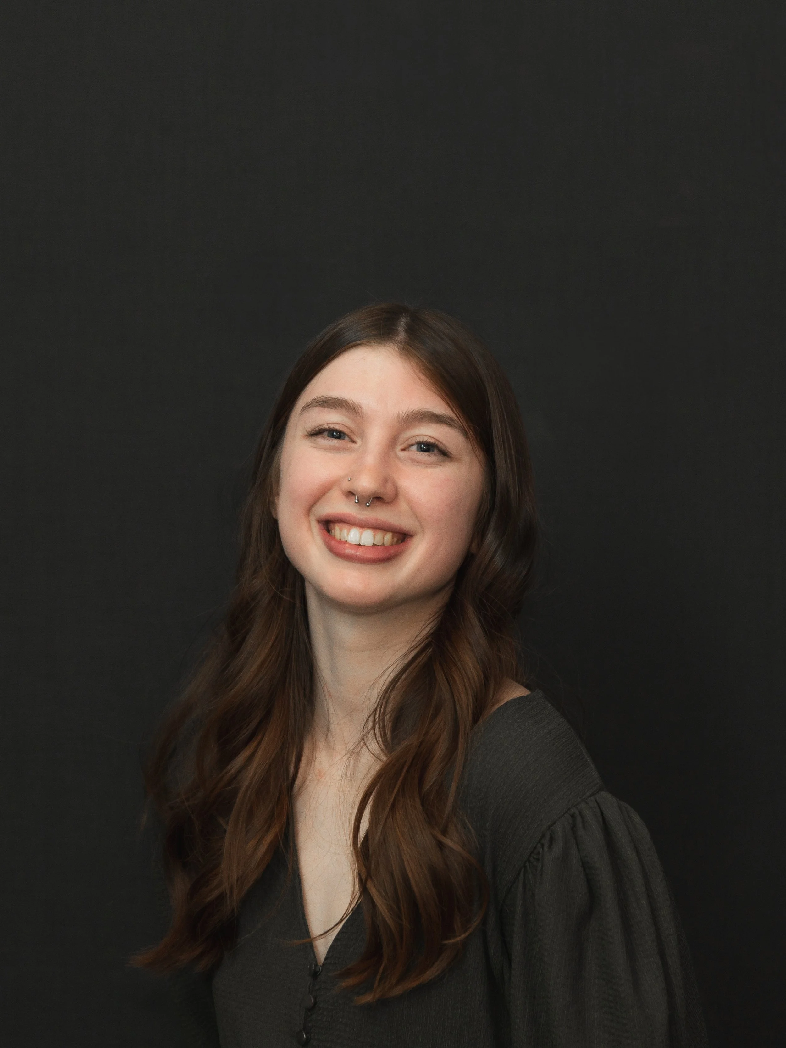 A young woman with long brown hair, smiling, wearing a black top, standing against a black background.