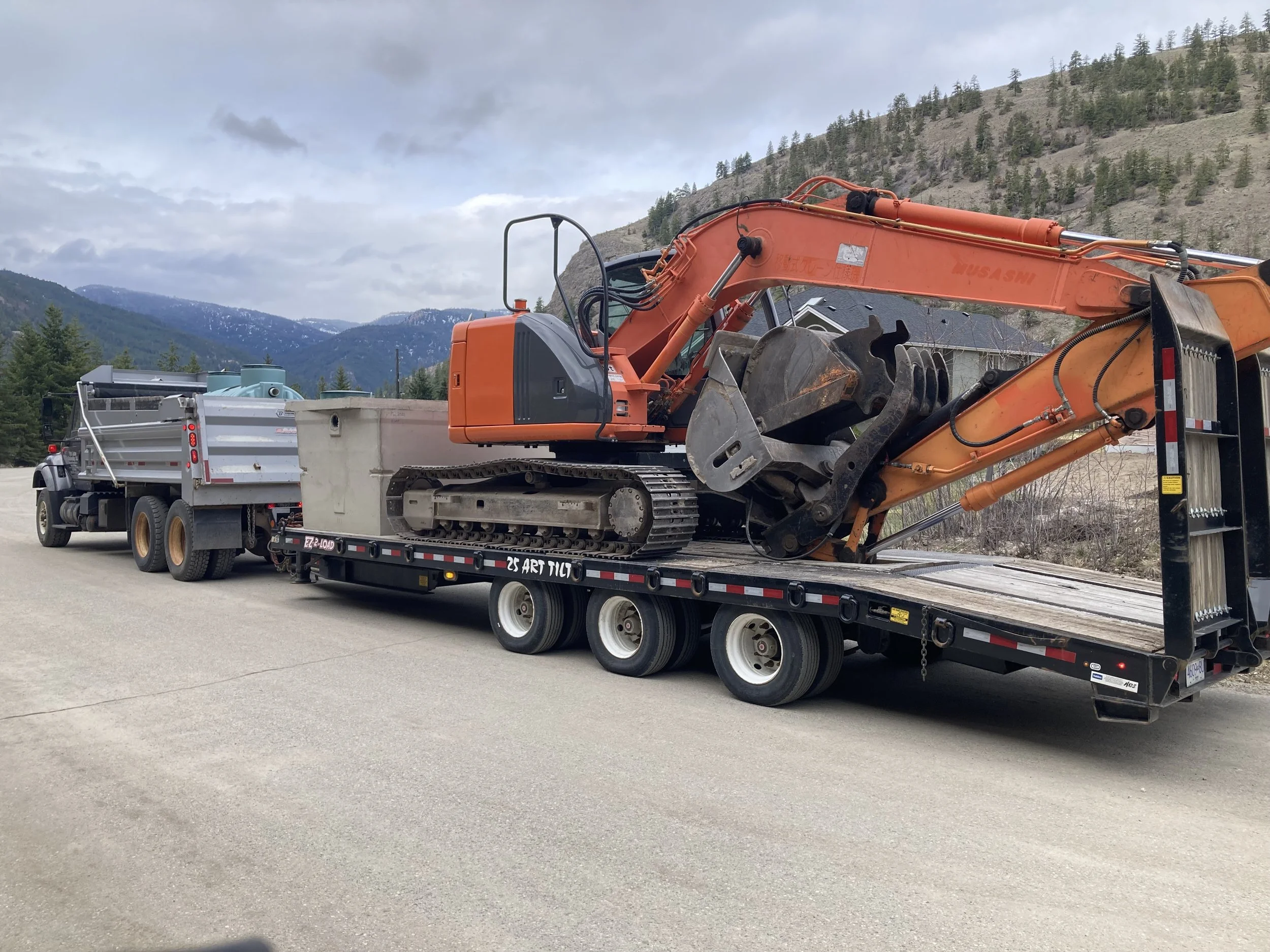 A large orange excavator is parked on a flatbed truck beside a paved road. The background features a mountainous landscape with scattered trees and cloudy skies.