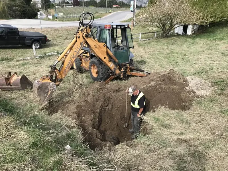 Rural property excavation project featuring a backhoe and worker beside a trench in the South Okanagan.