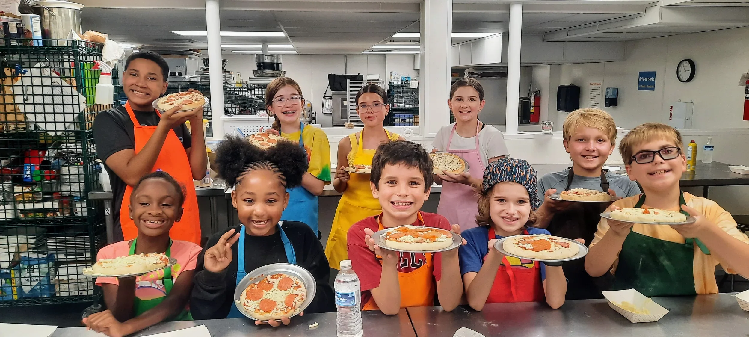 Several campers holding their pizzas they made during camp and smiling before they bake them with their aprons on.