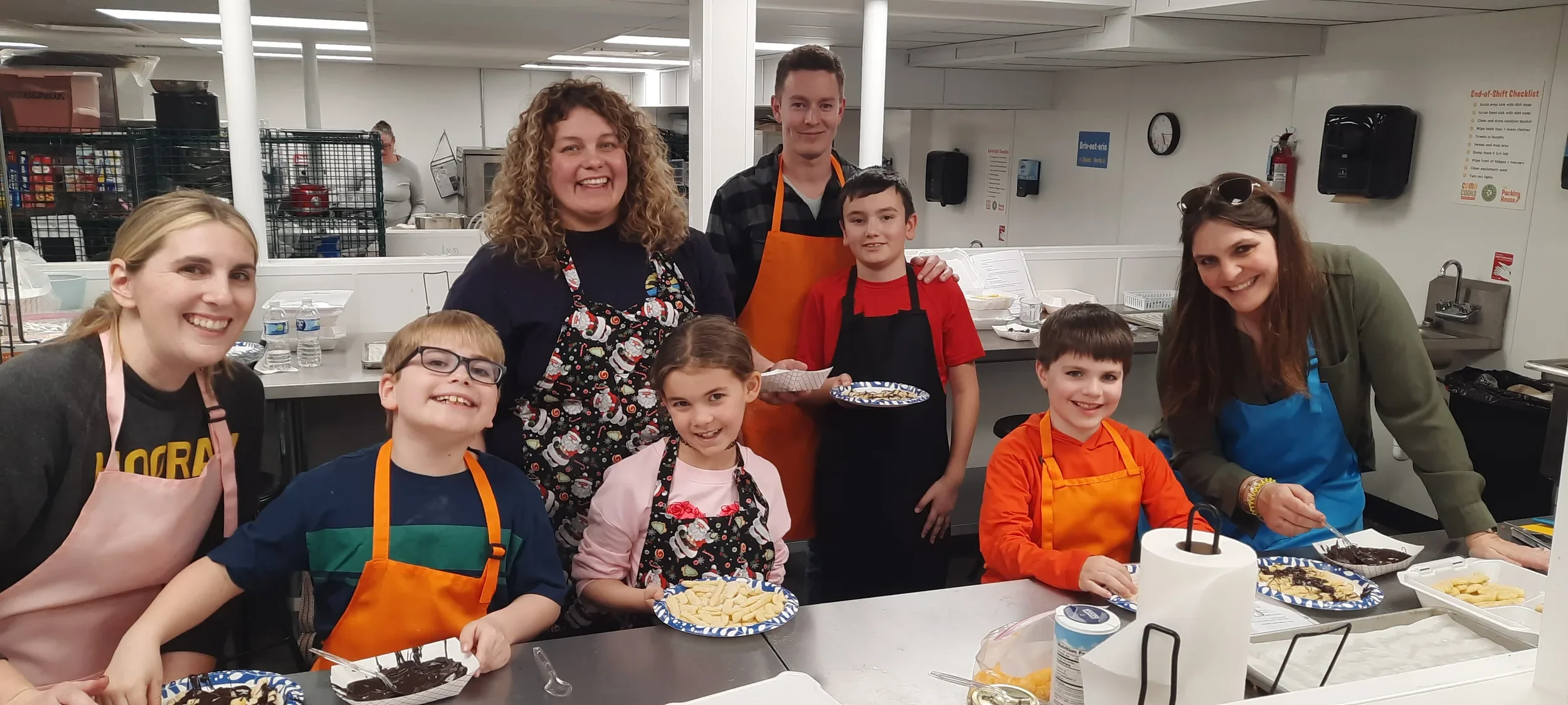 A group of parents and their kids (4 of each) holding their dishes they made during their class before eating all with smiles on their faces and aprons on in the kitchen