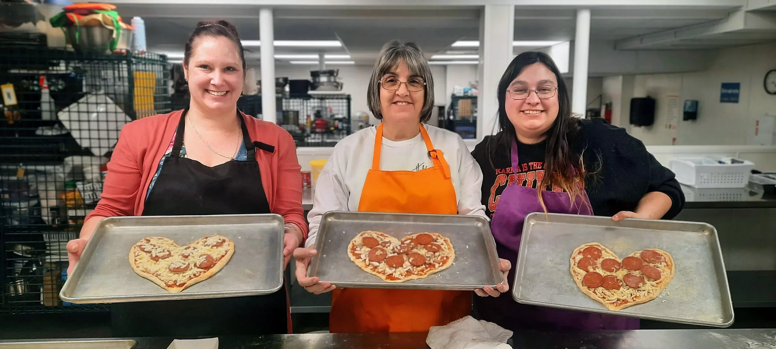 Three students wearing their aprons and holding their sheet pans with their heart shaped pizzas ready to bake with smiles on their faces