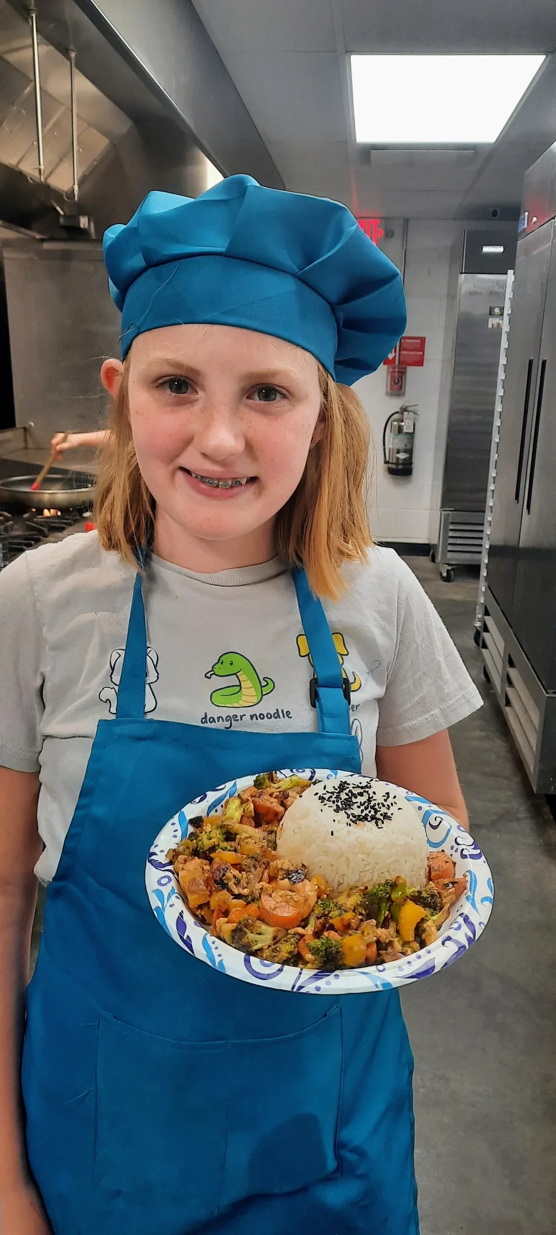Teen in a blue apron and chef hat holding her beautifully plated stir fry she created without a recipe during cooking camp