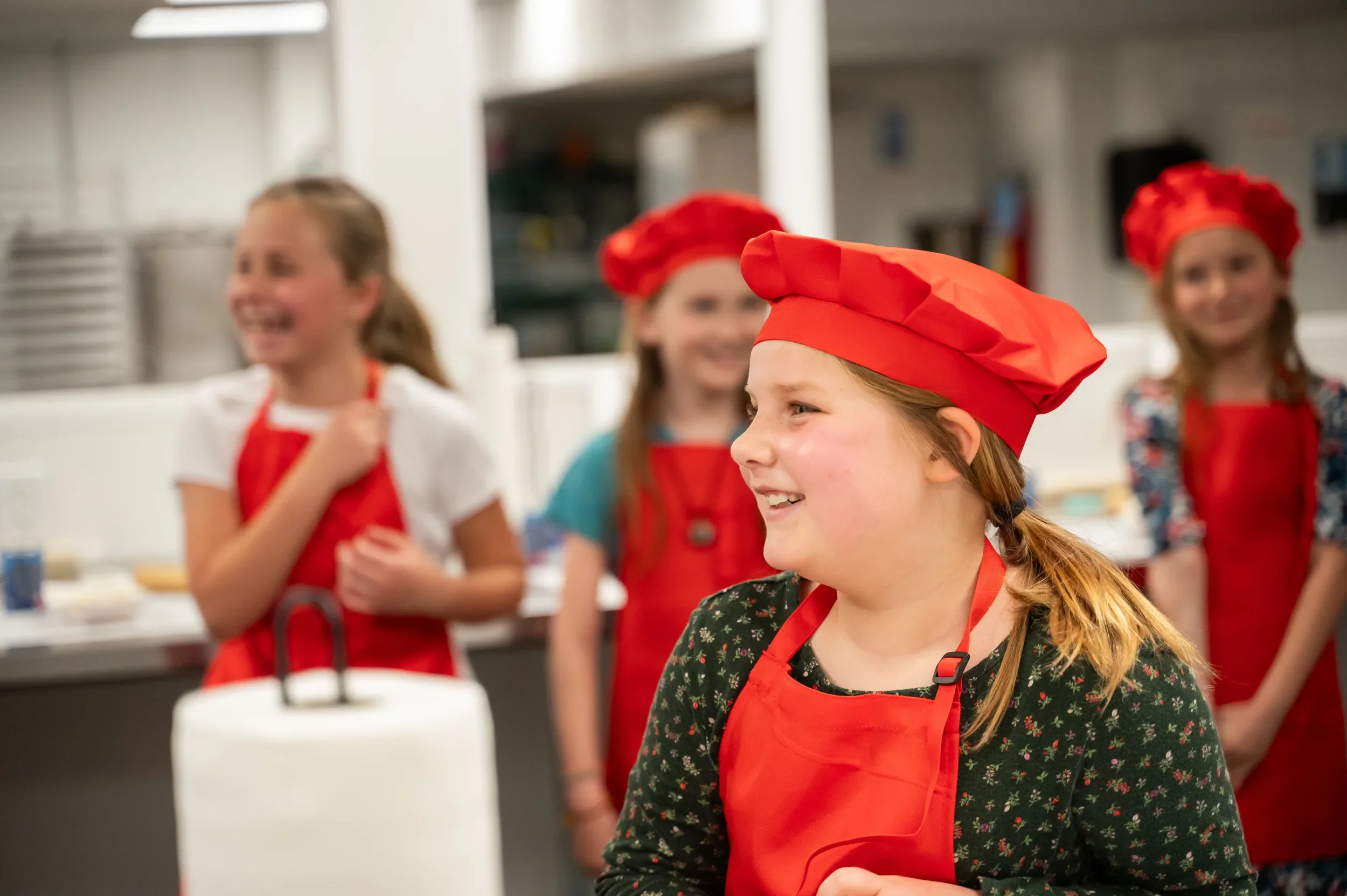 Smiling girl in a red chef hat and apron at a kids cooking class, other young chefs behind her