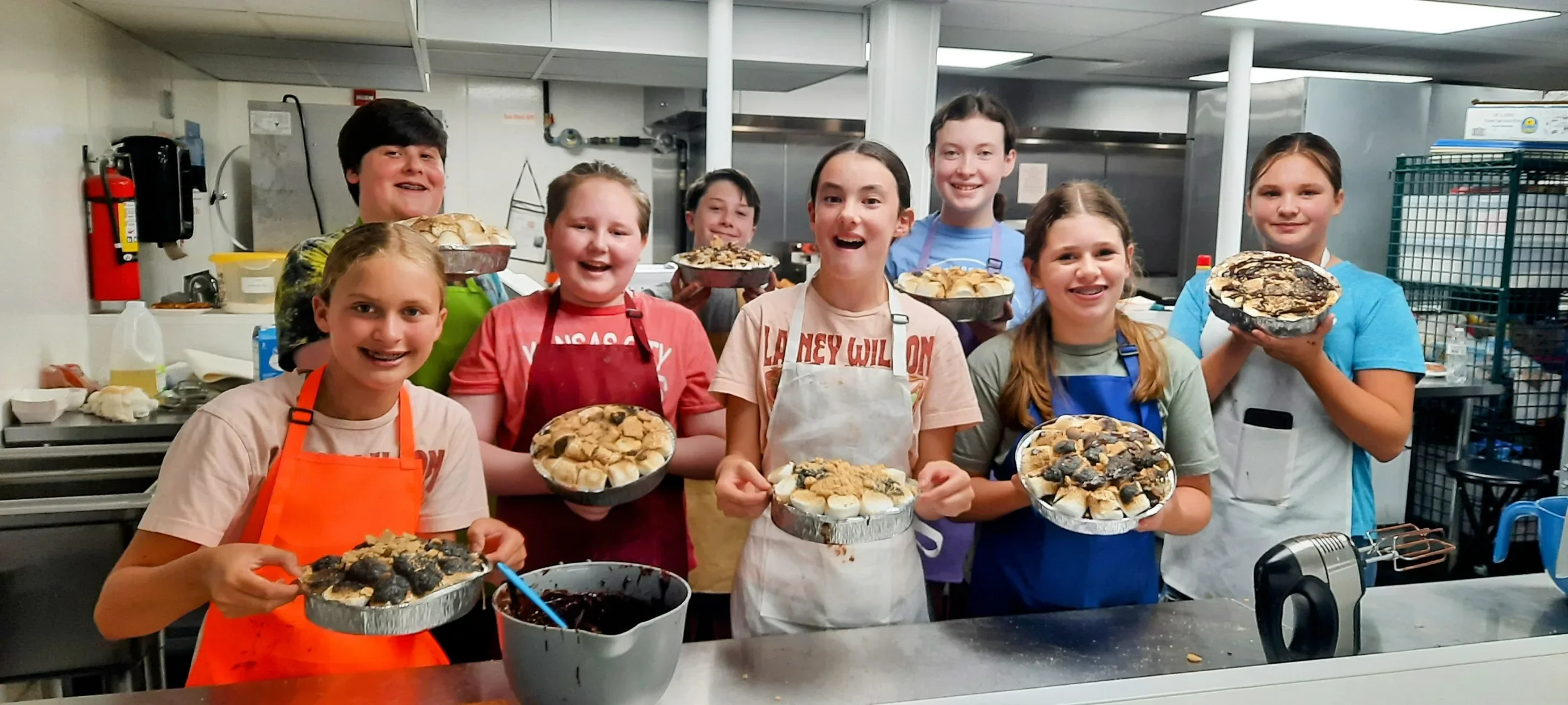 Several happy teens holding their s'mores cakes they made during camp with smiles on their faces and their colorful aprons on.