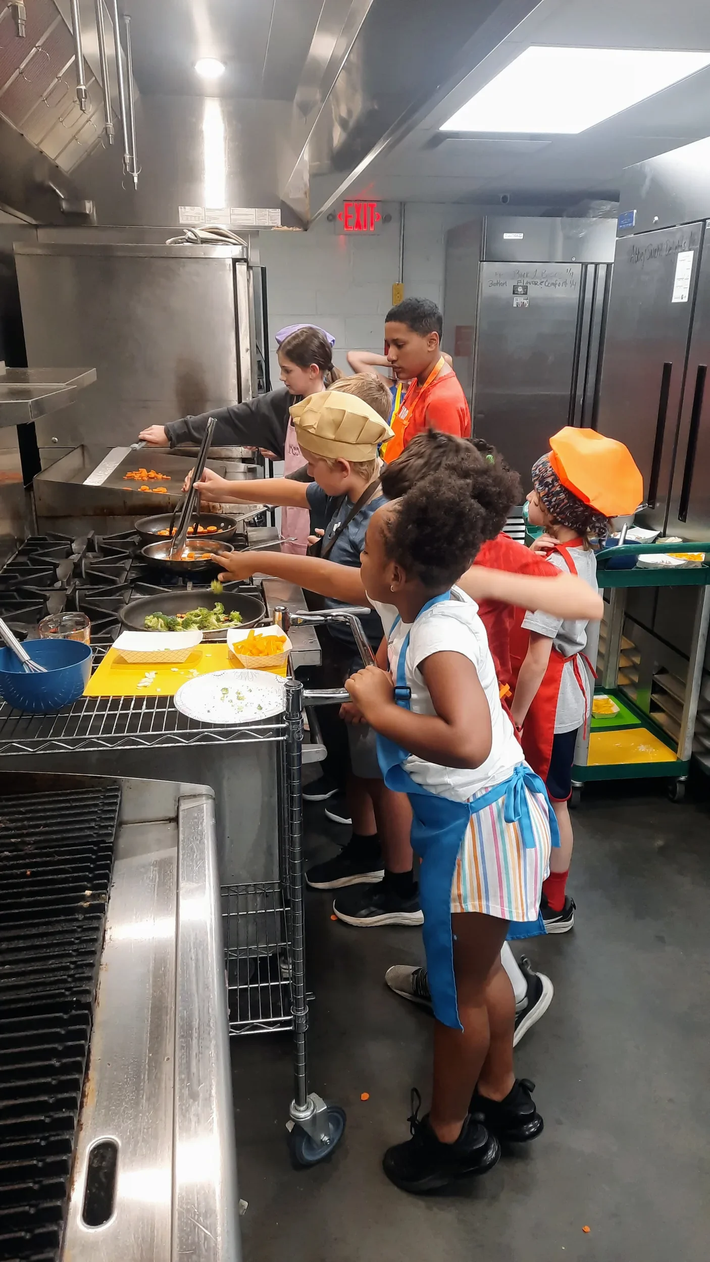 Kids in aprons sautéing carrots and broccoli for stir-fry at the stove during a cooking camp