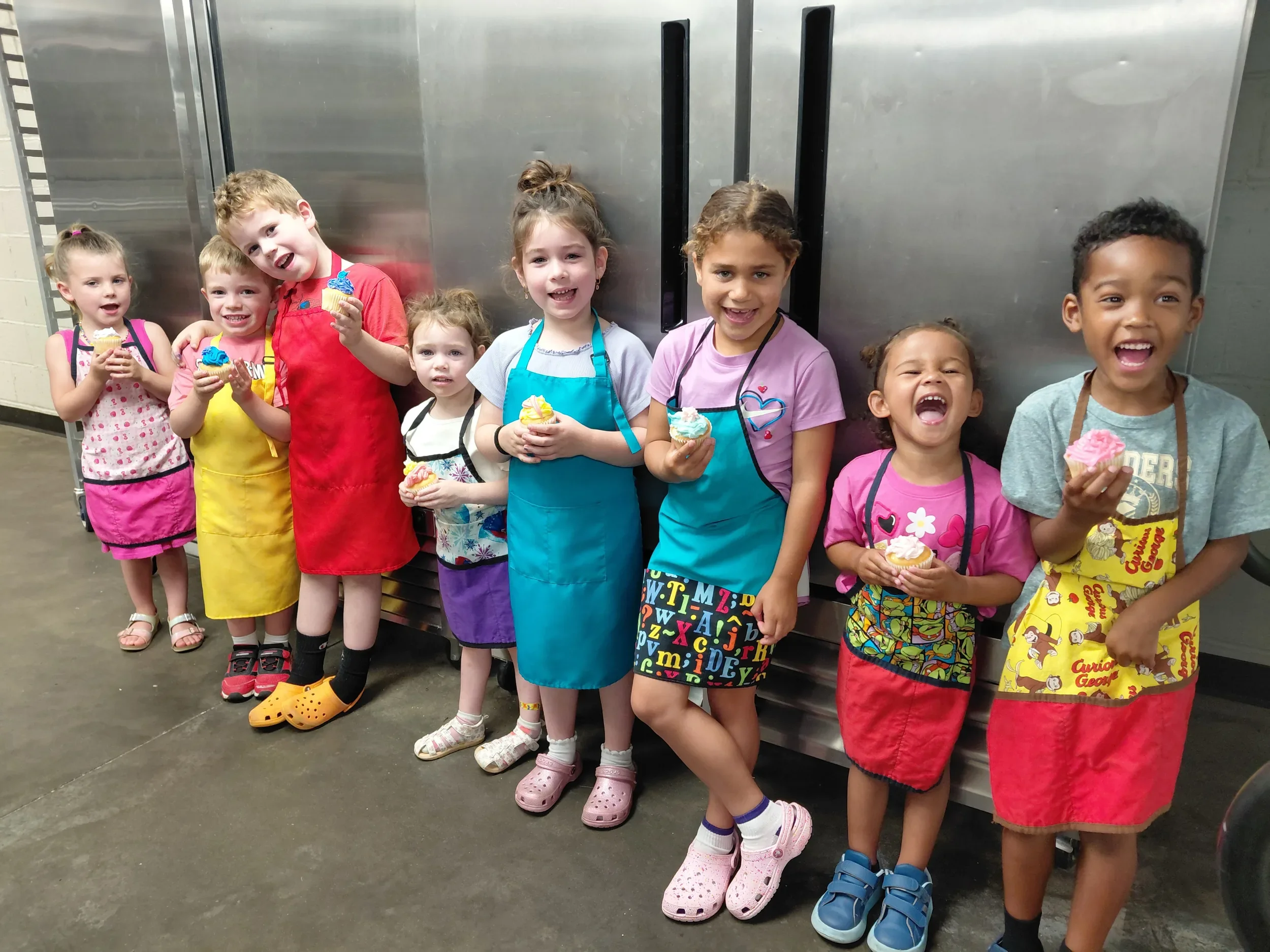 Our youngest chefs ages 3-6 years old in aprons grinning and holding the cupcakes they frosted in a Mini Chef class.