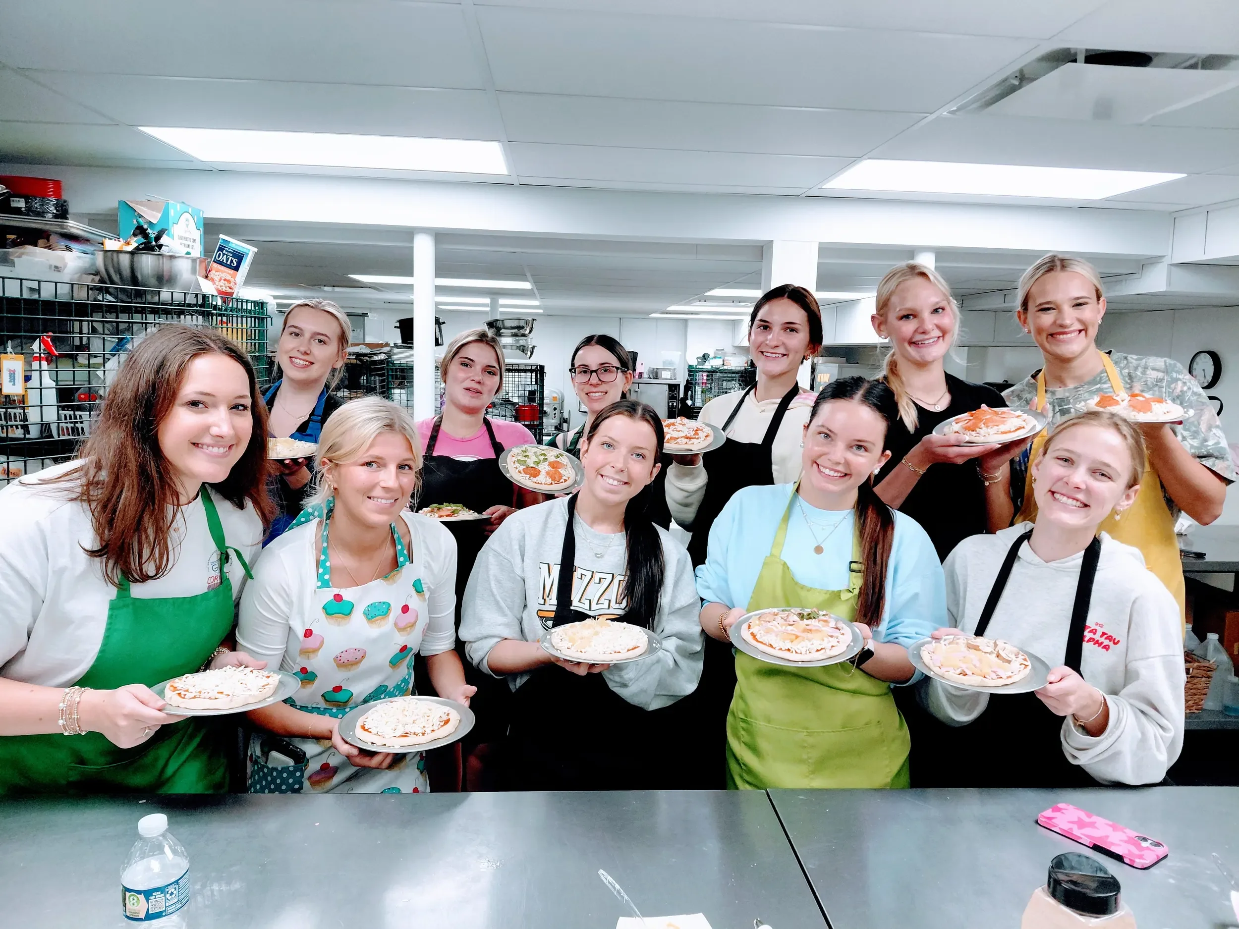 Large group of female students with aprons on holding their freshly made personal pizzas they made during their cooking class while smiling