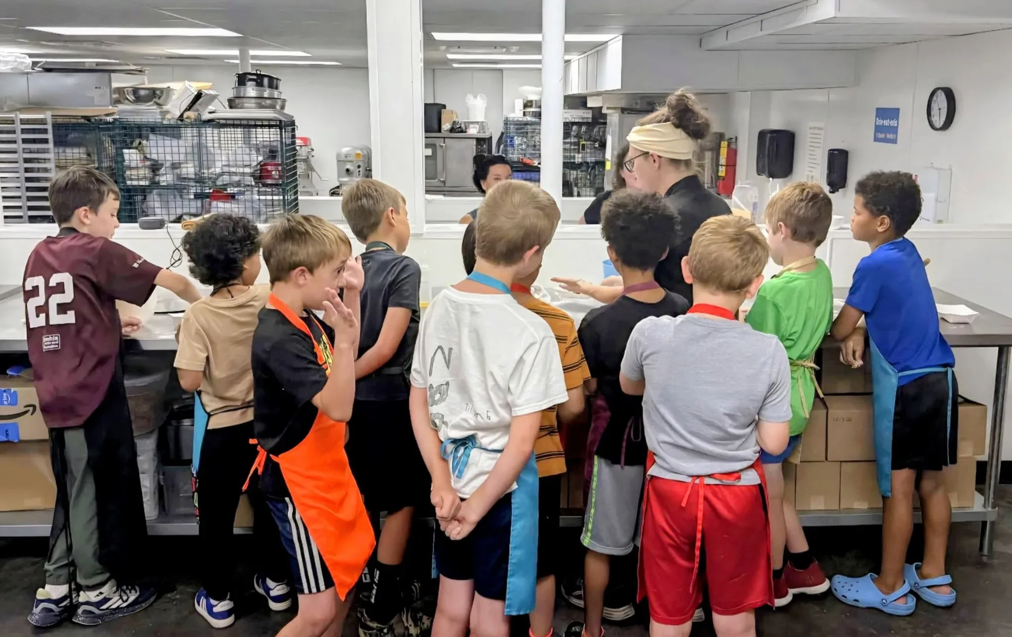 A group of kids gathered around Chef Ashley at a kitchen counter during a cooking class