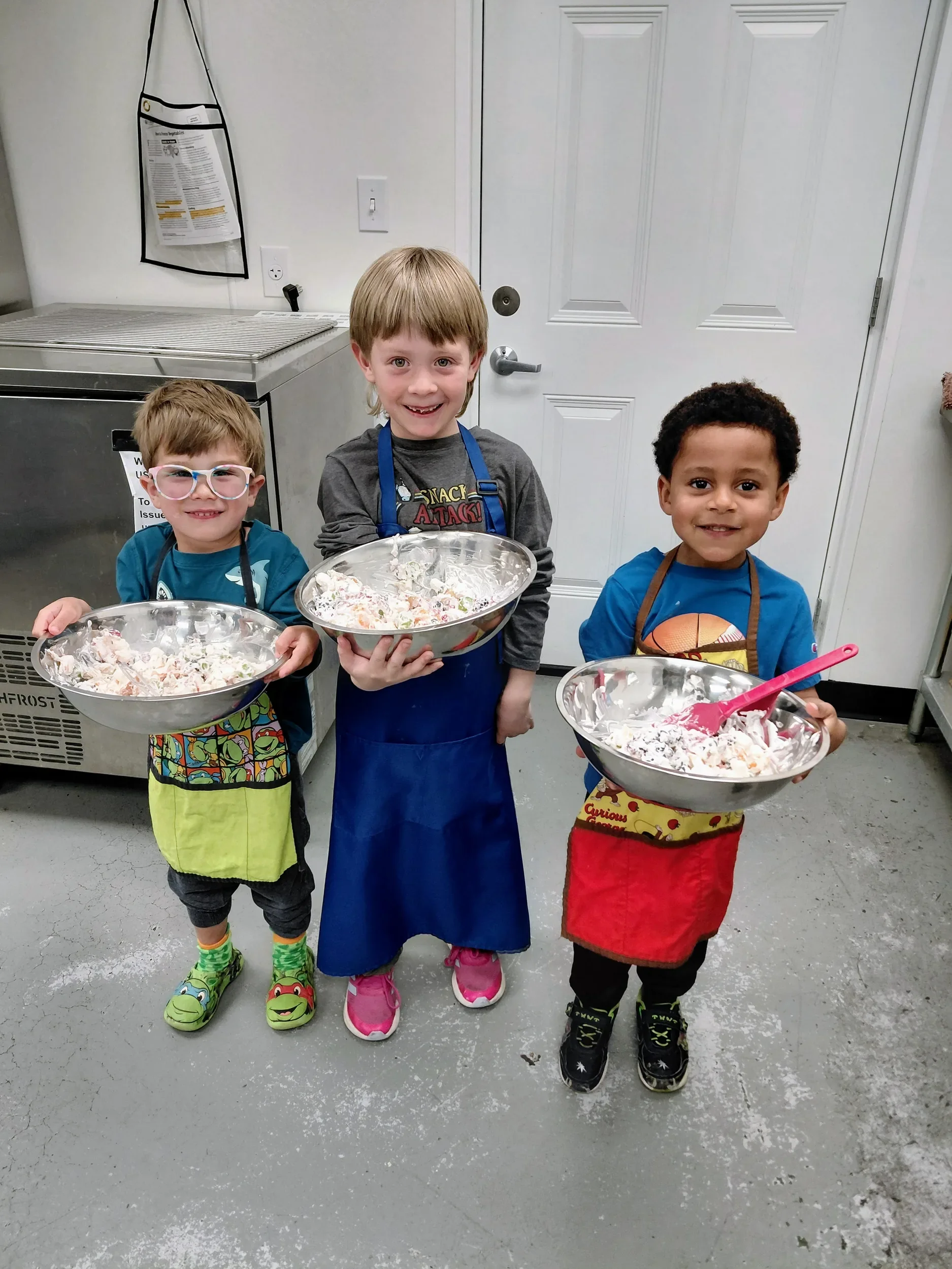 Three mini chefs ages 3-6 years old proudly holding the bowls of fruit salad they mixed.