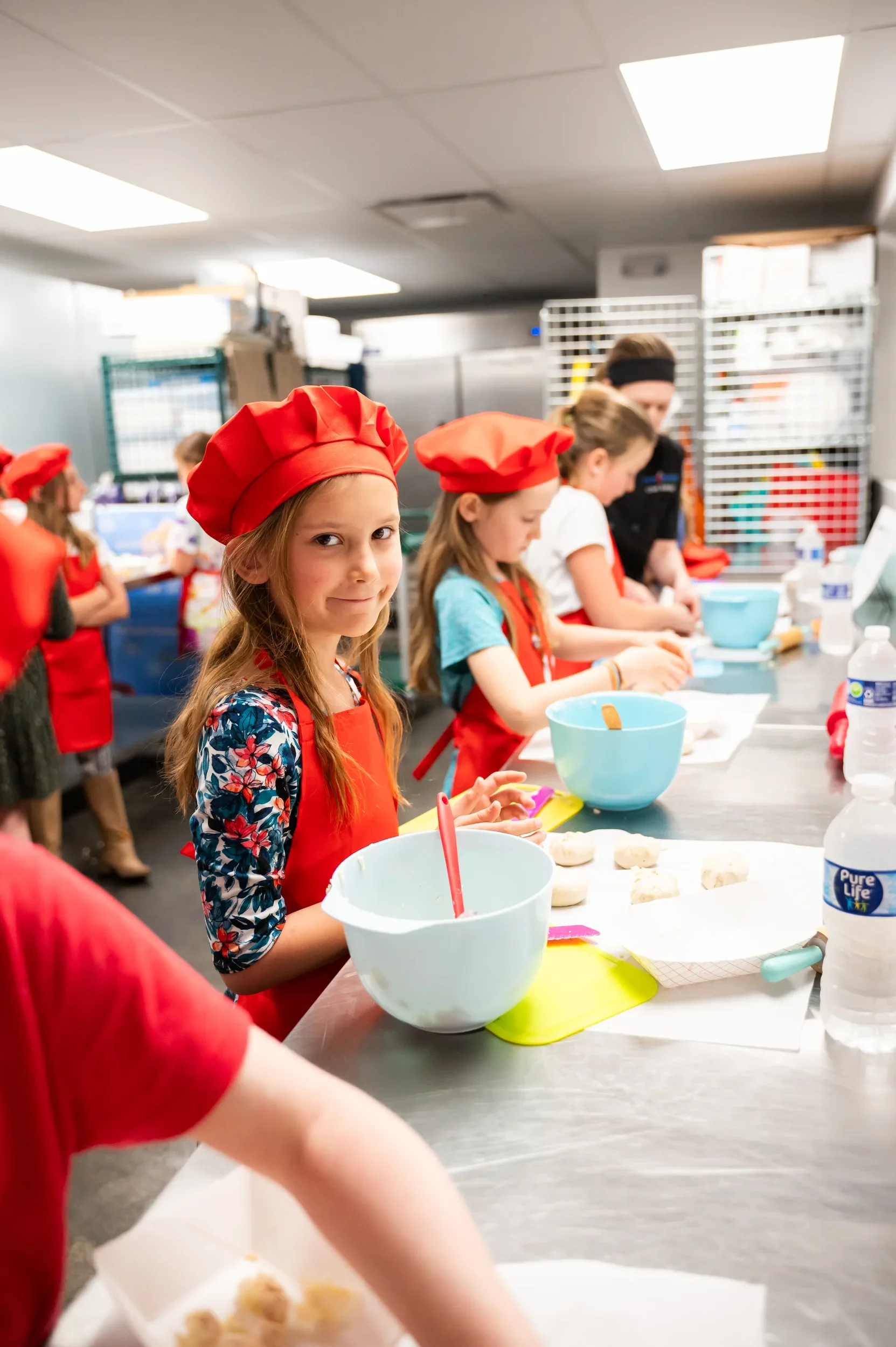 Children in red chef hats and red aprons working in their stations to prepare their cookies during their class while Chef Ashley is shown helping one of the students in the background.