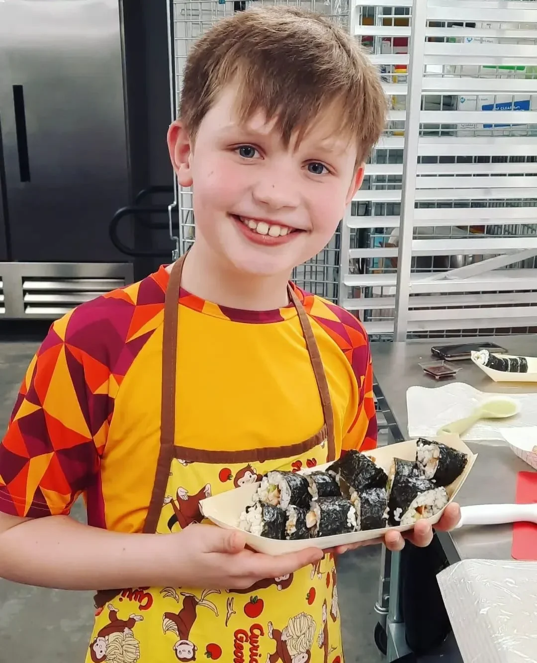 Boy in his apron holding the sushi he just made during a sushi class. He has a smile on his face.