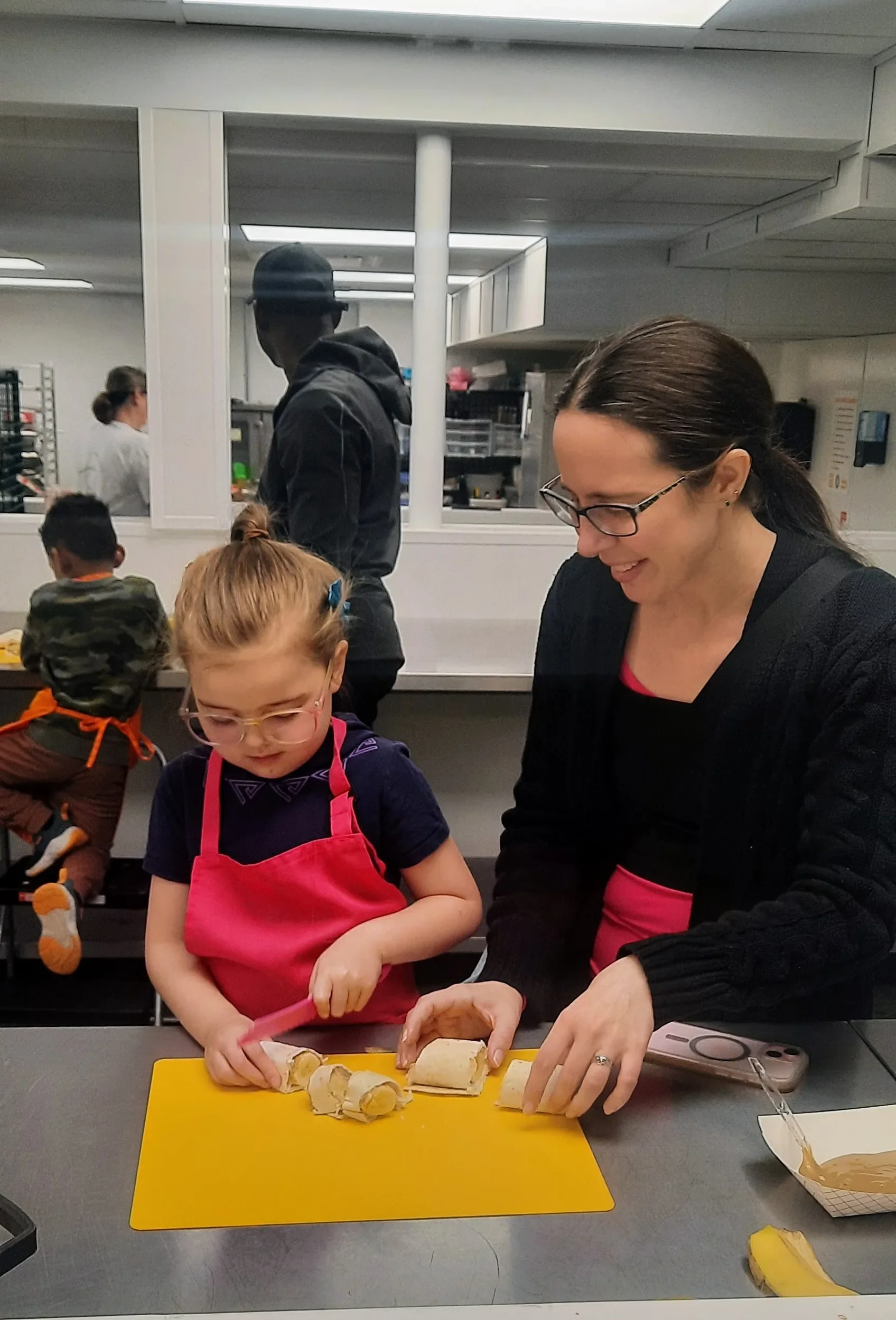 Young child slicing a peanut butter banana roll-up with a parent helping during a mini chef cooking class