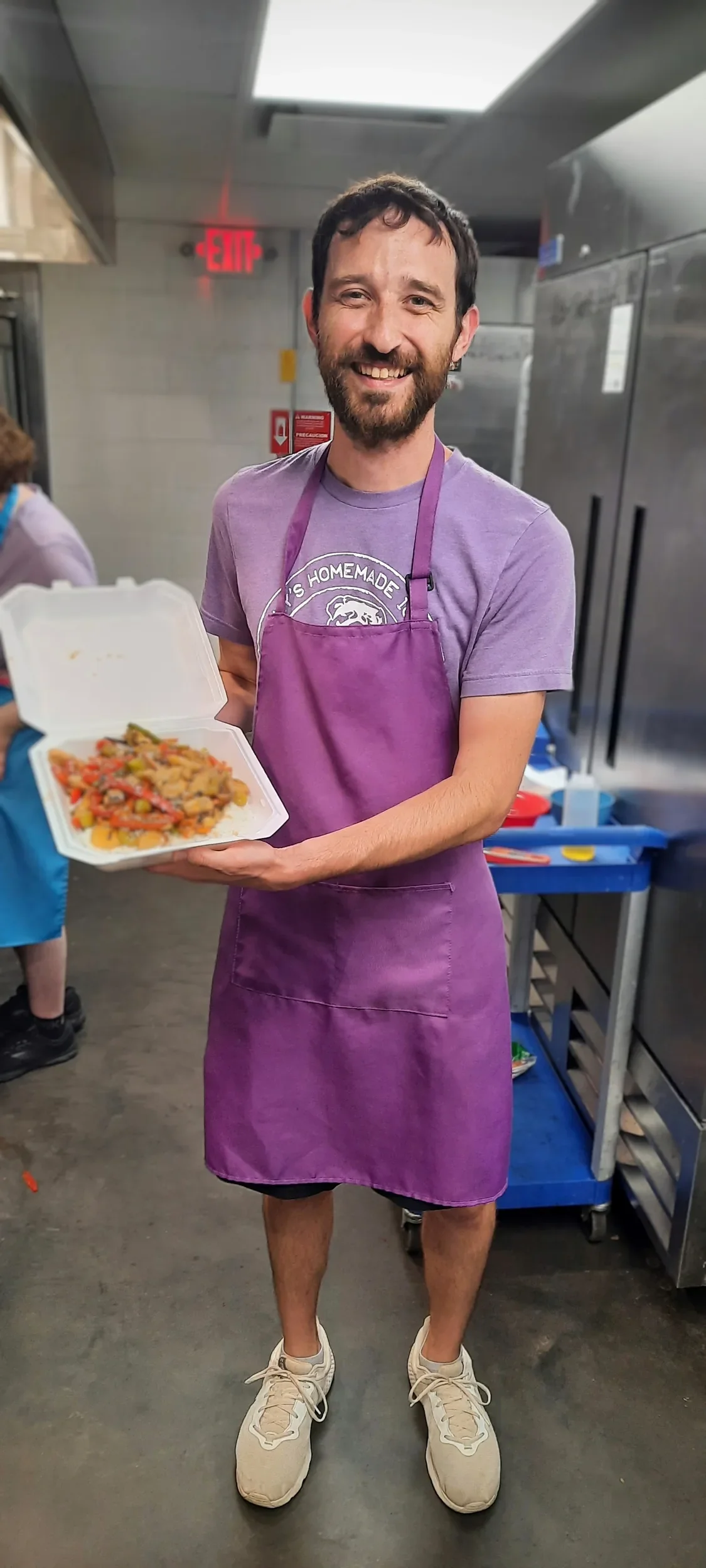 Student in purple apron and a smile on his face holding up the stir-fry he prepared during the knife skills class from the cooks in the making series
