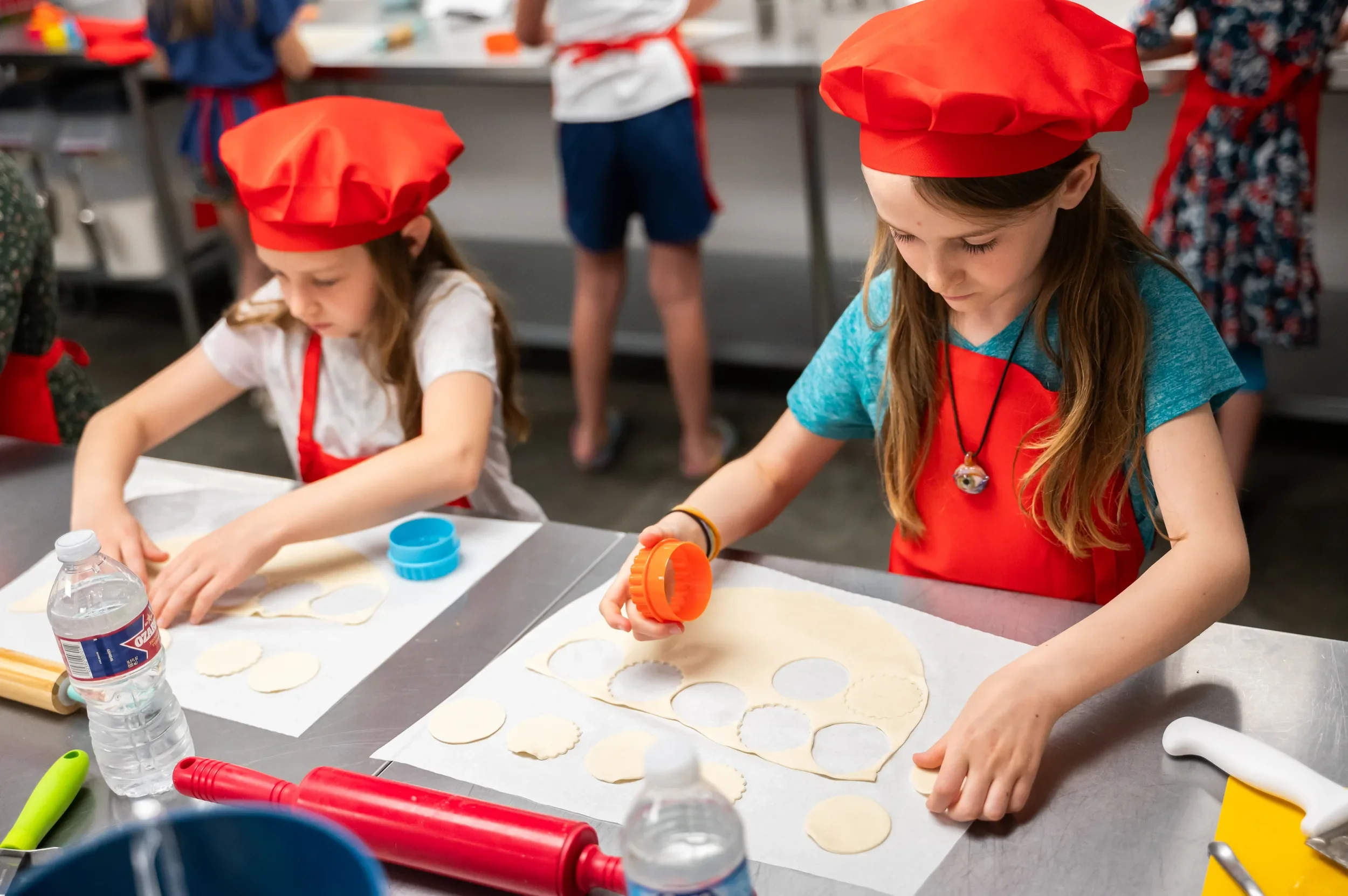 Two girls in red chef hats and aprons cutting dough rounds with cutters for apple pie cookies at a kids cooking class