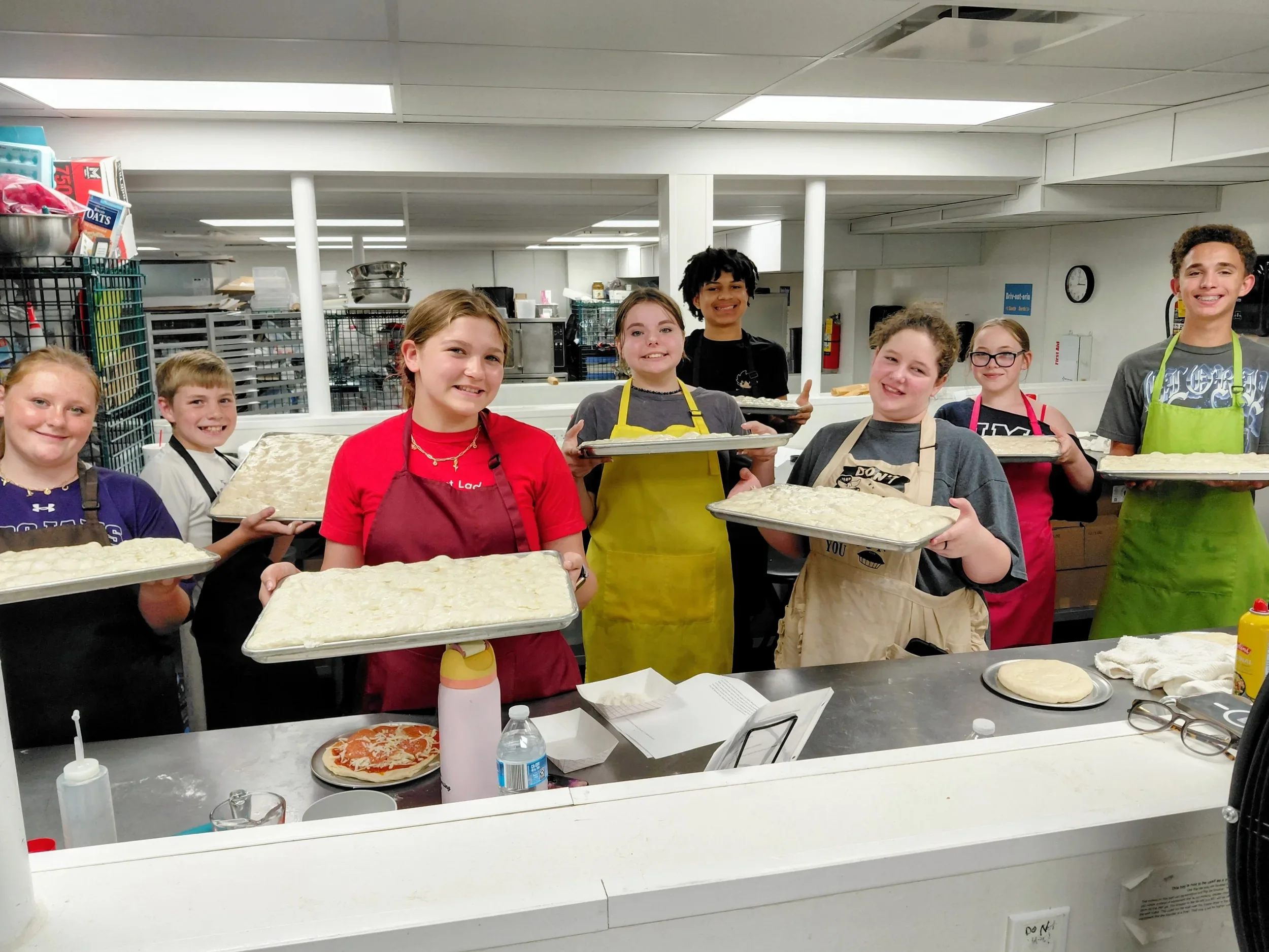 Teens with their aprons on and dough on a tray with mostly smiles on their faces.