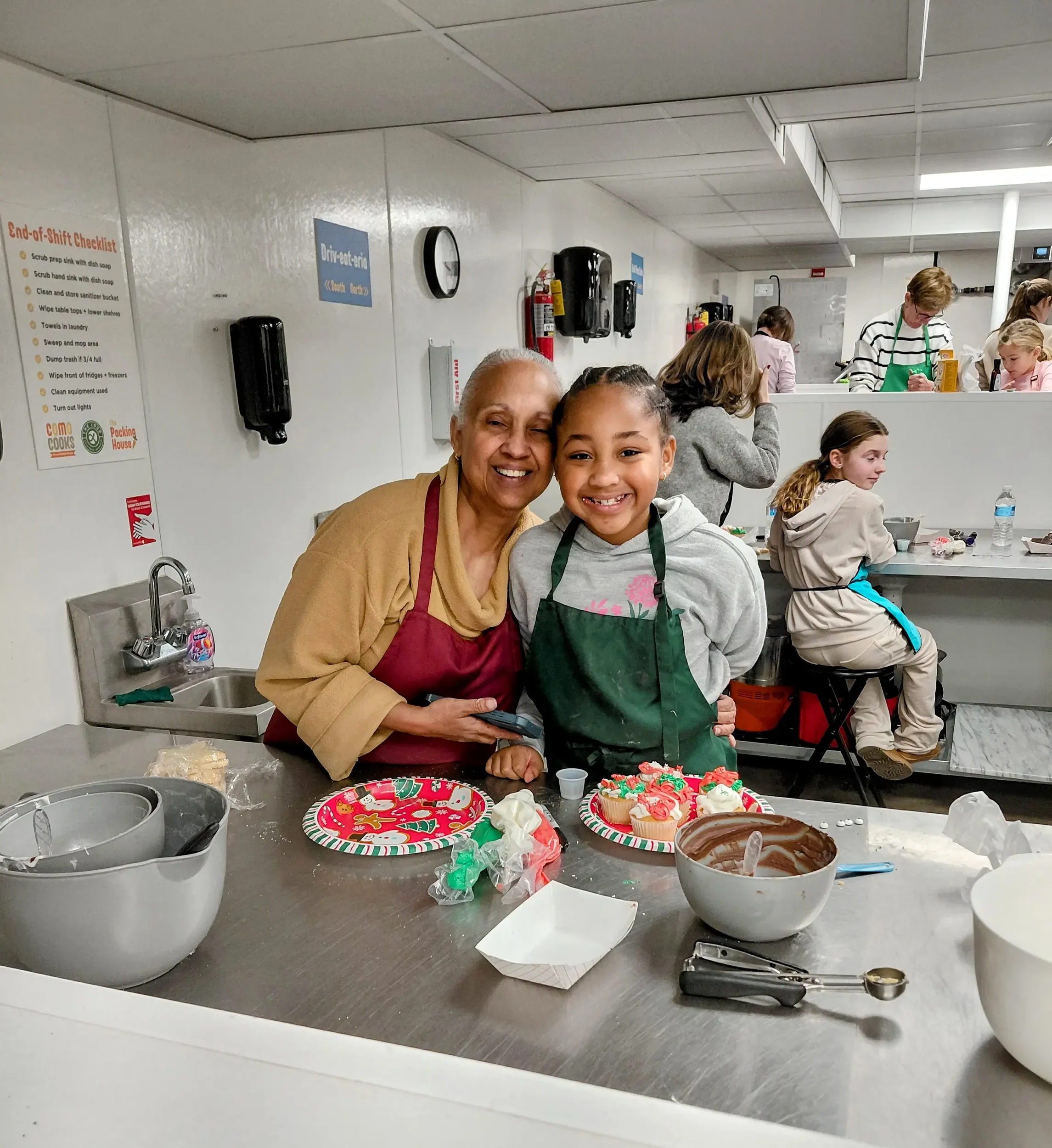 A grandmother and granddaughter smiling with their aprons on while standing in front of their decorated cupcakes during a holiday treats workshop