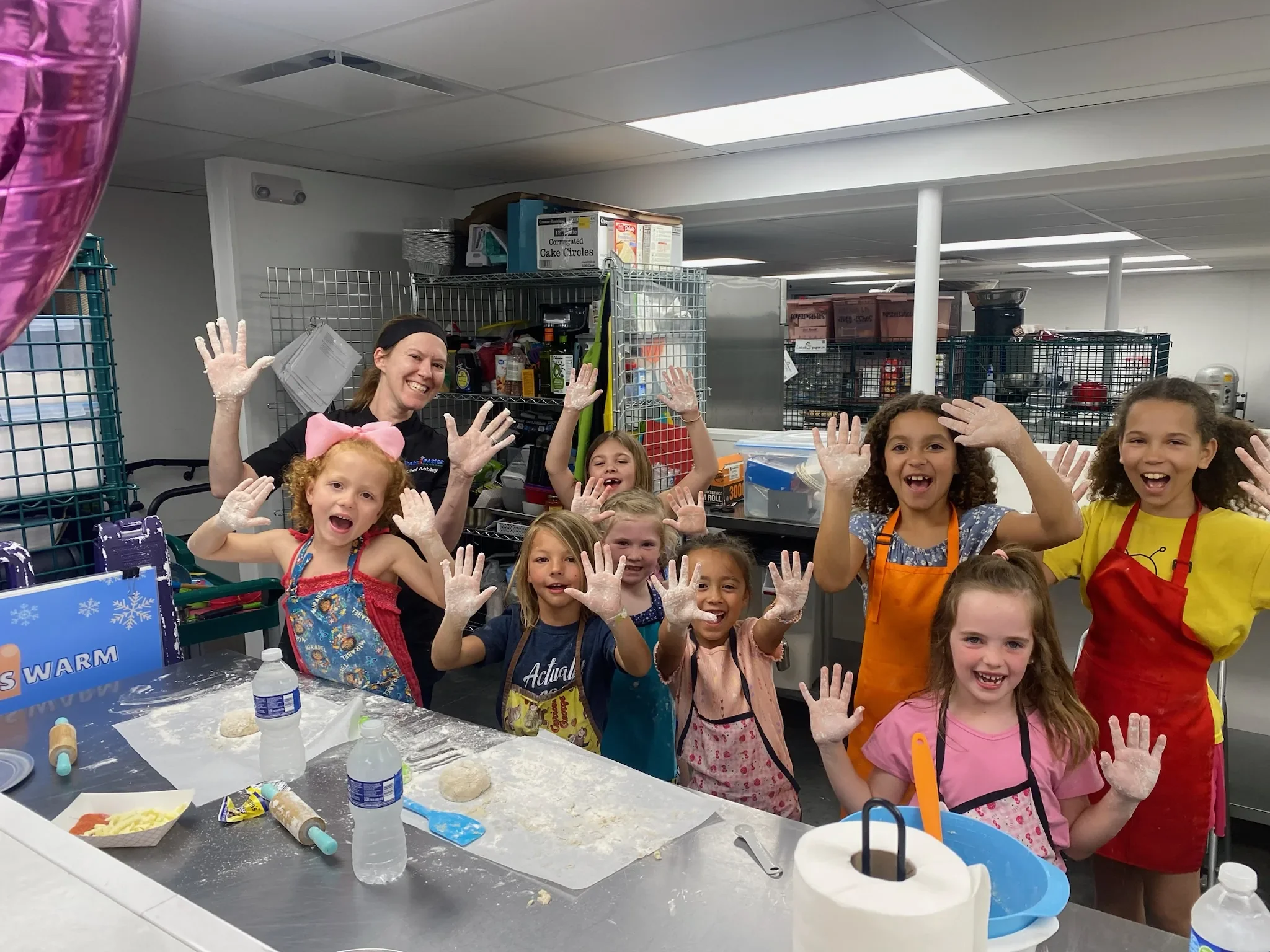 Chef Ashley and a group of children holding up their flour covered hands with smiles and aprons on during their cooking class