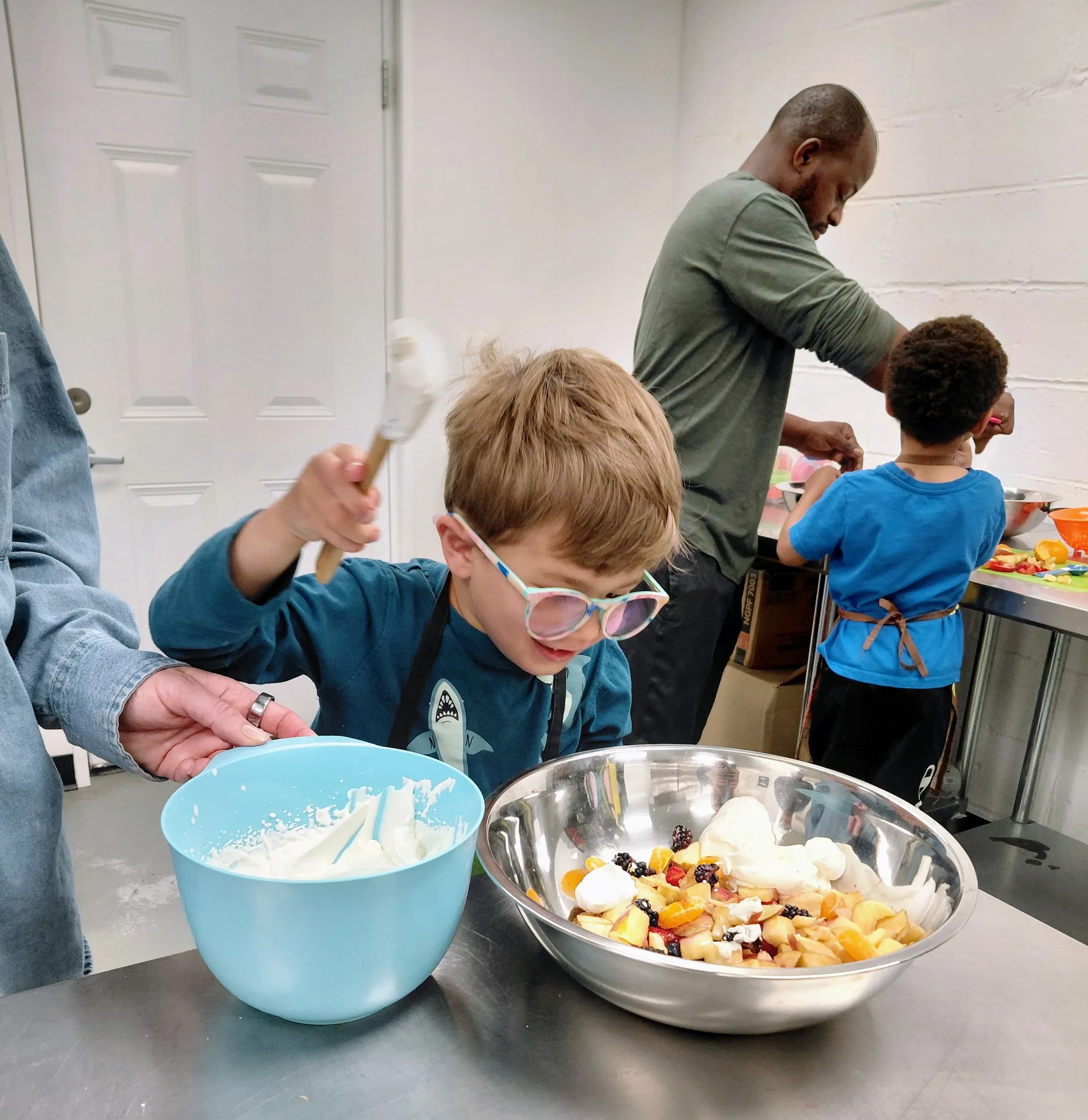 Boy in glasses whisking cream while an adult steadies his bowl, a dad and child cooking behind during a mini chef class