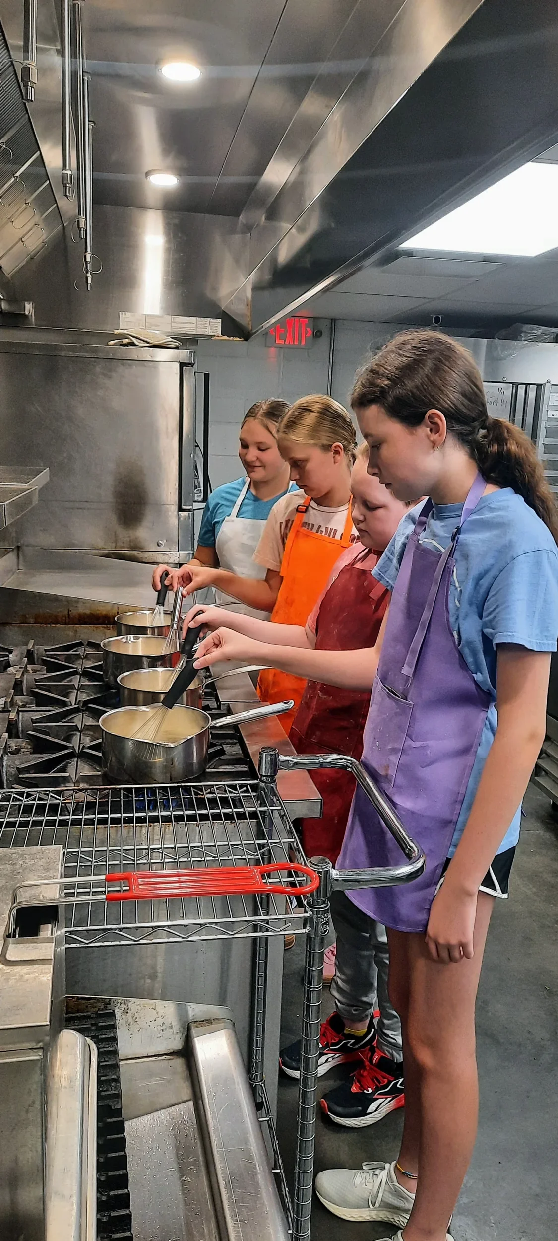 Four teens at the stovetop mixing their sauce with a whisk in their pan with different colored aprons one during class.