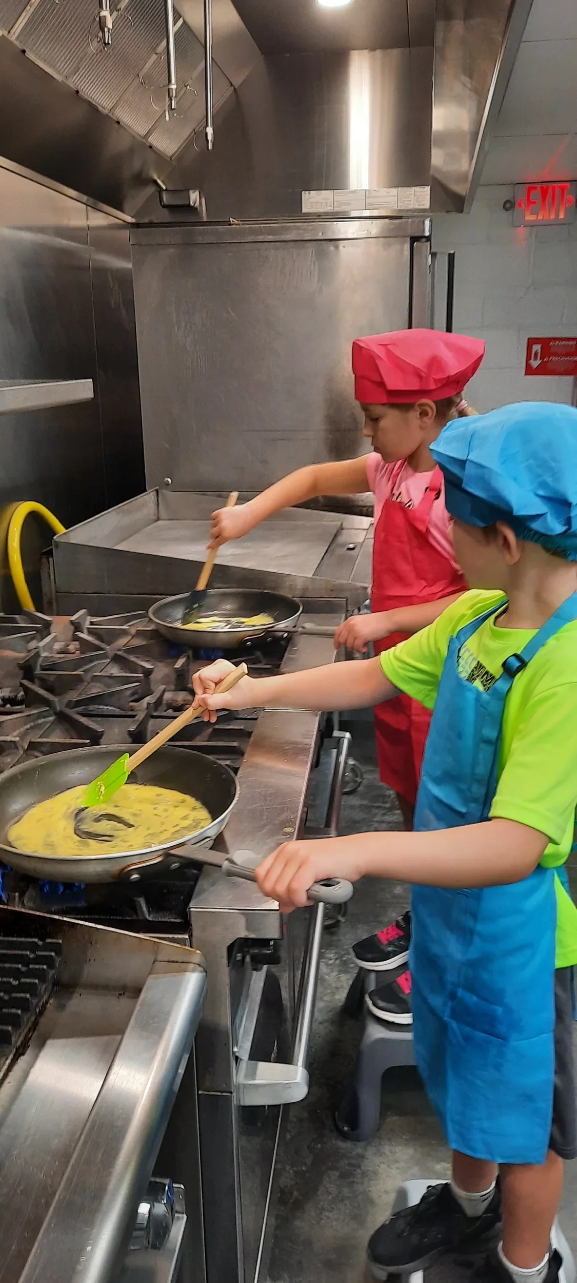 Two children one in blue and one in pink aprons and chef hat cooking scrambled eggs during cooking camp