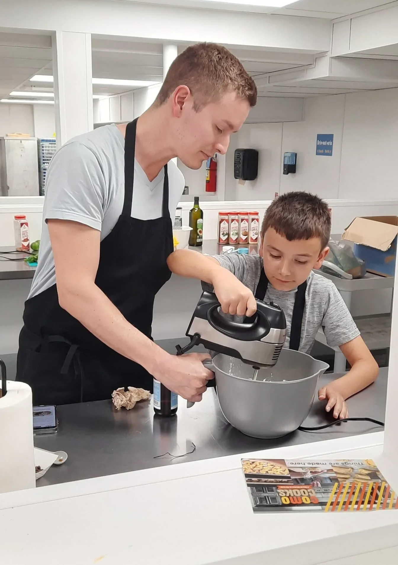 Father holding a mix bowl peeking down at the bowl while his son holds a hand mixer and mixes their ingredients during class