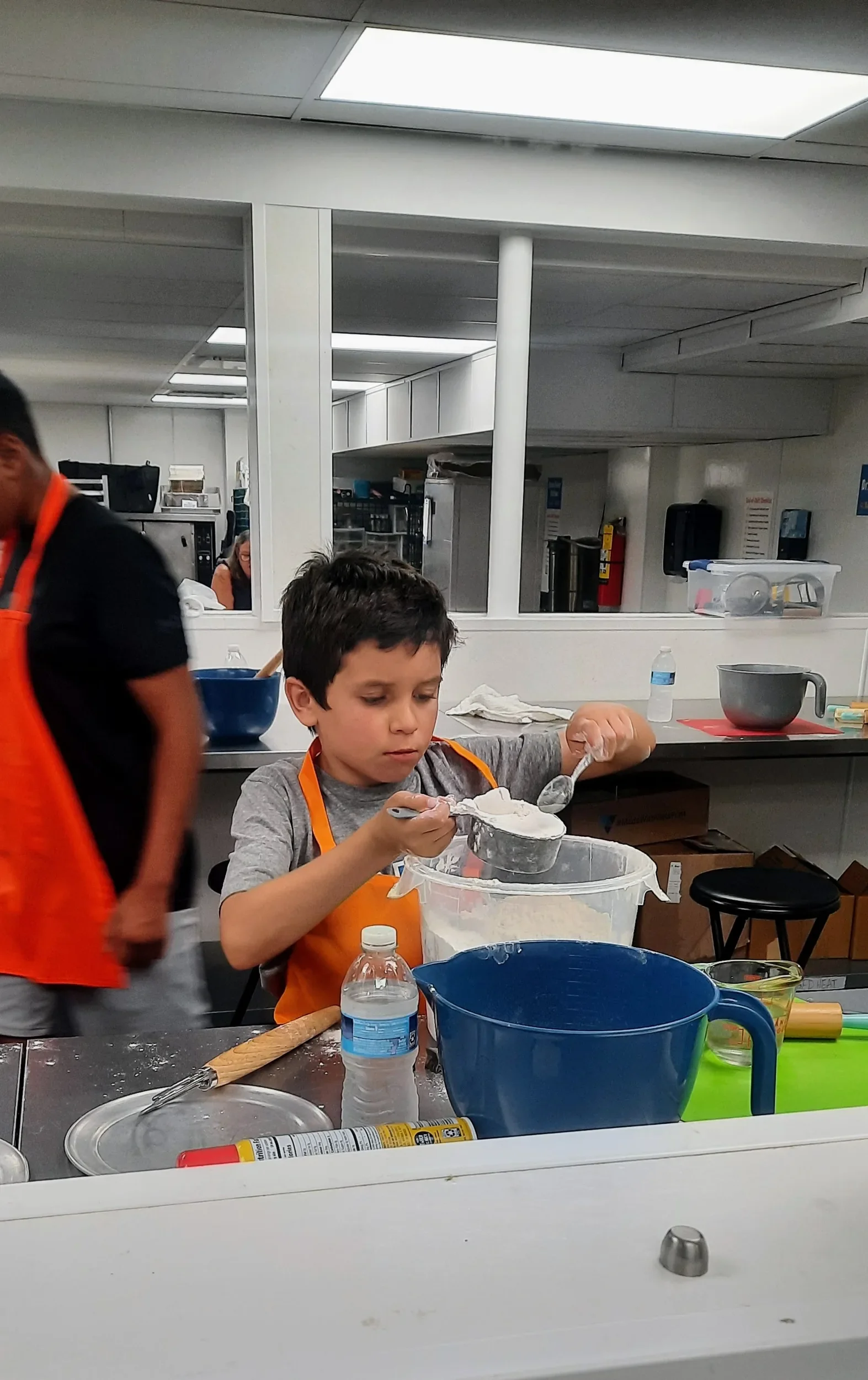 Young camper with a bright orange apron on, carefully measuring and leveling flour into a measuring cup while baking
