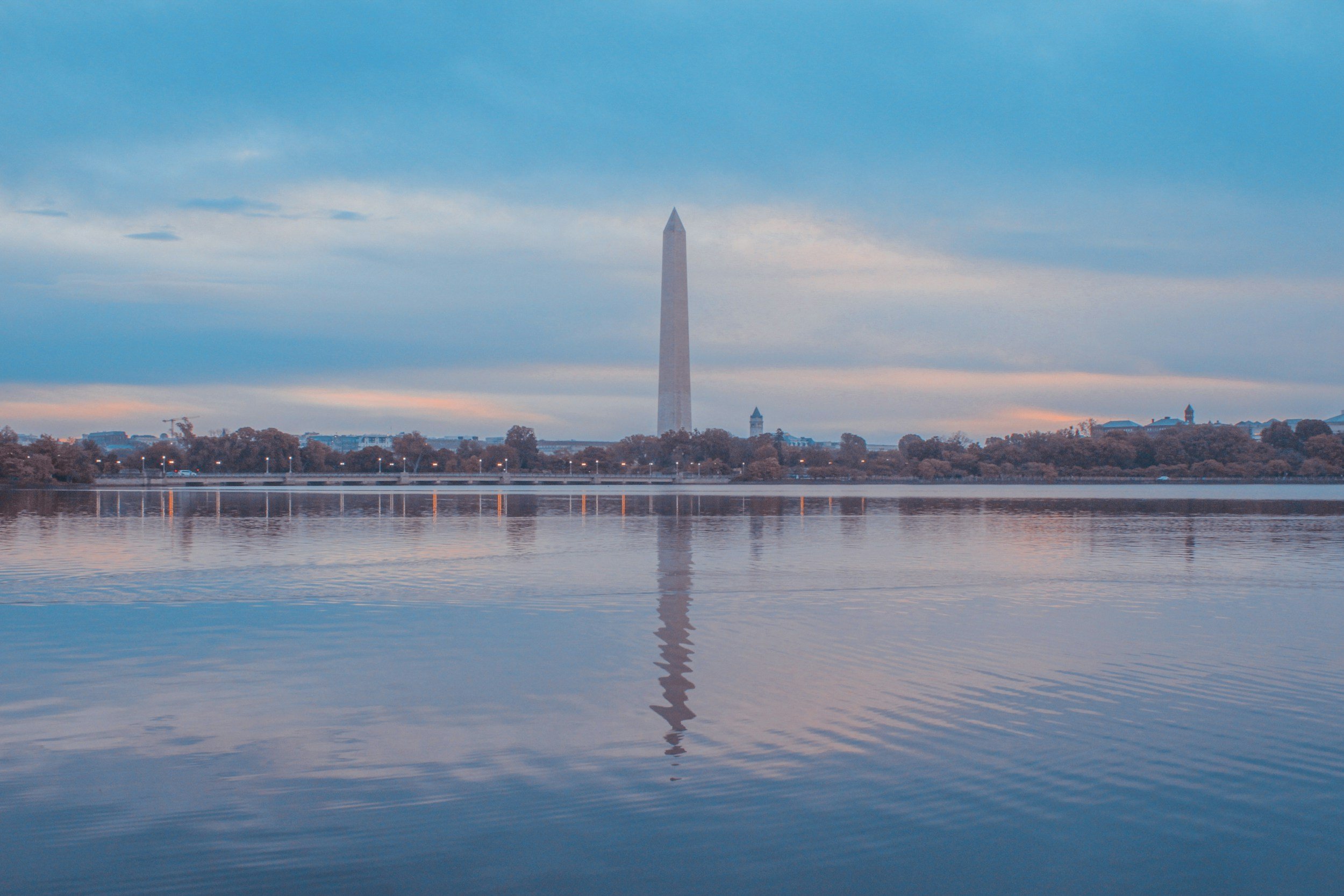 The Washington Monument seen across a body of water during sunset, with a partly cloudy sky and trees in the foreground.