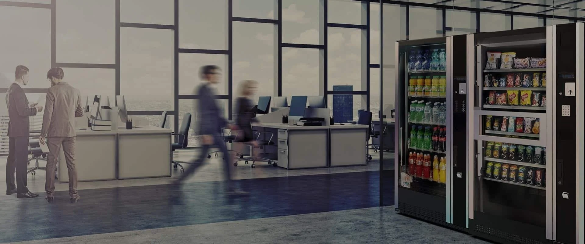 Modern office space with two vending machines filled with drinks, three people walking past desks with computers, and two people in business attire talking near a desk during daytime.