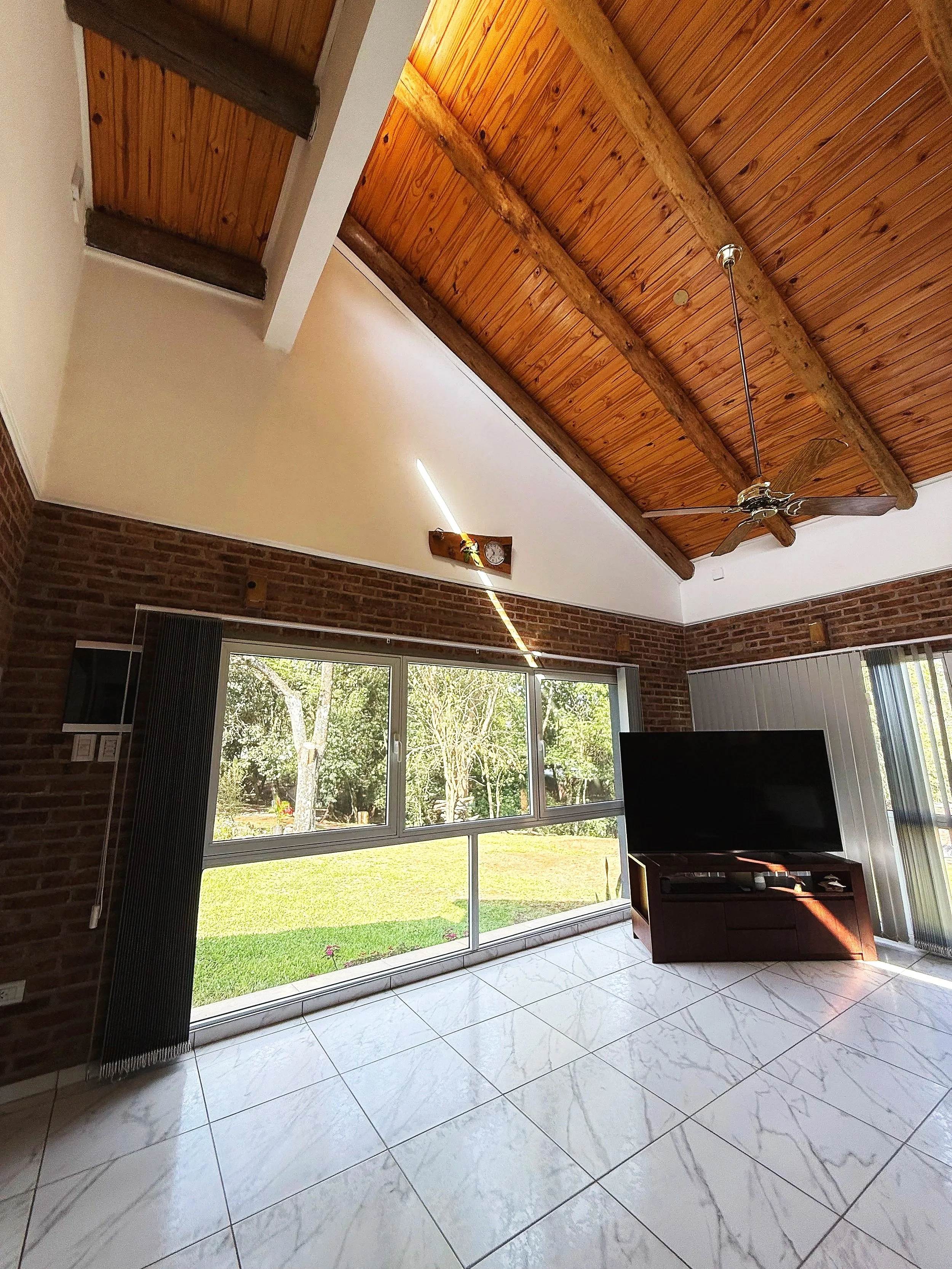 Interior de una sala con ventanas grandes que muestran un jardín verde, techo de madera con vigas y ventilador, televisión y suelo de baldosas blancas con vetas grises.