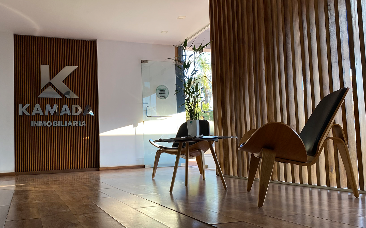 Interior de oficina inmobiliaria con una pared de madera, dos sillas de diseño moderno, una mesa con papeles y un folder, y un logo de 'KAMADA INMOBILIARIA' en la pared.