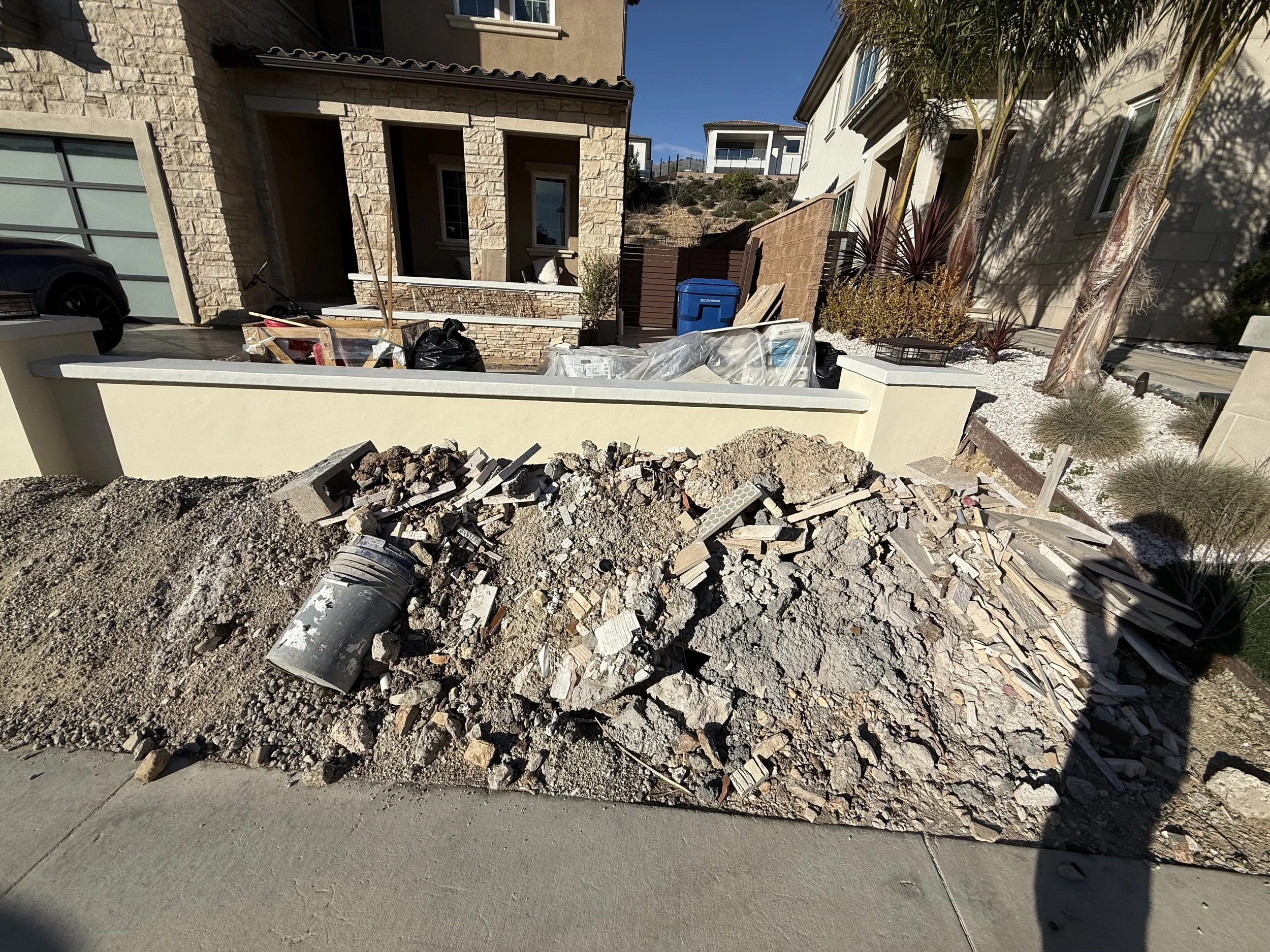 Construction site in a residential neighborhood with a pile of gravel, bricks, and construction materials in the foreground, and houses with trees and landscaping in the background.