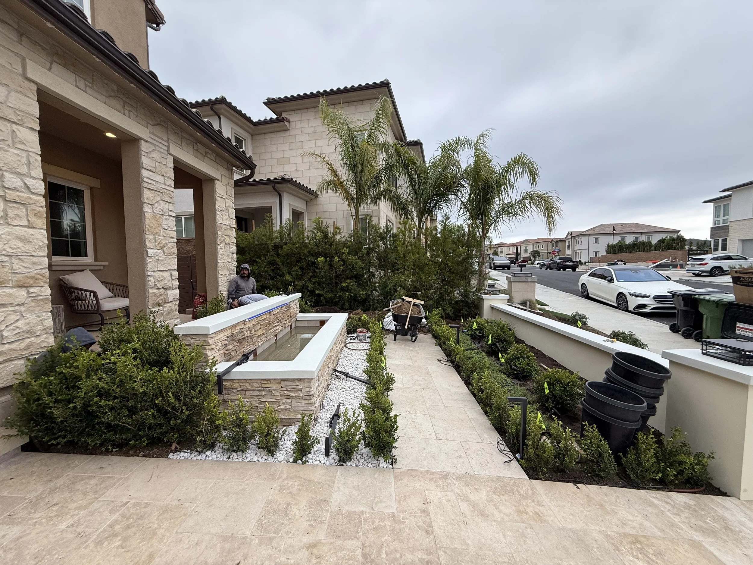 A suburban residential street with modern houses, cars parked along the street, and a partly cloudy sky. In the foreground, a house has a patio with a man sitting on a porch swing, surrounded by a landscaped garden with small bushes and trees.