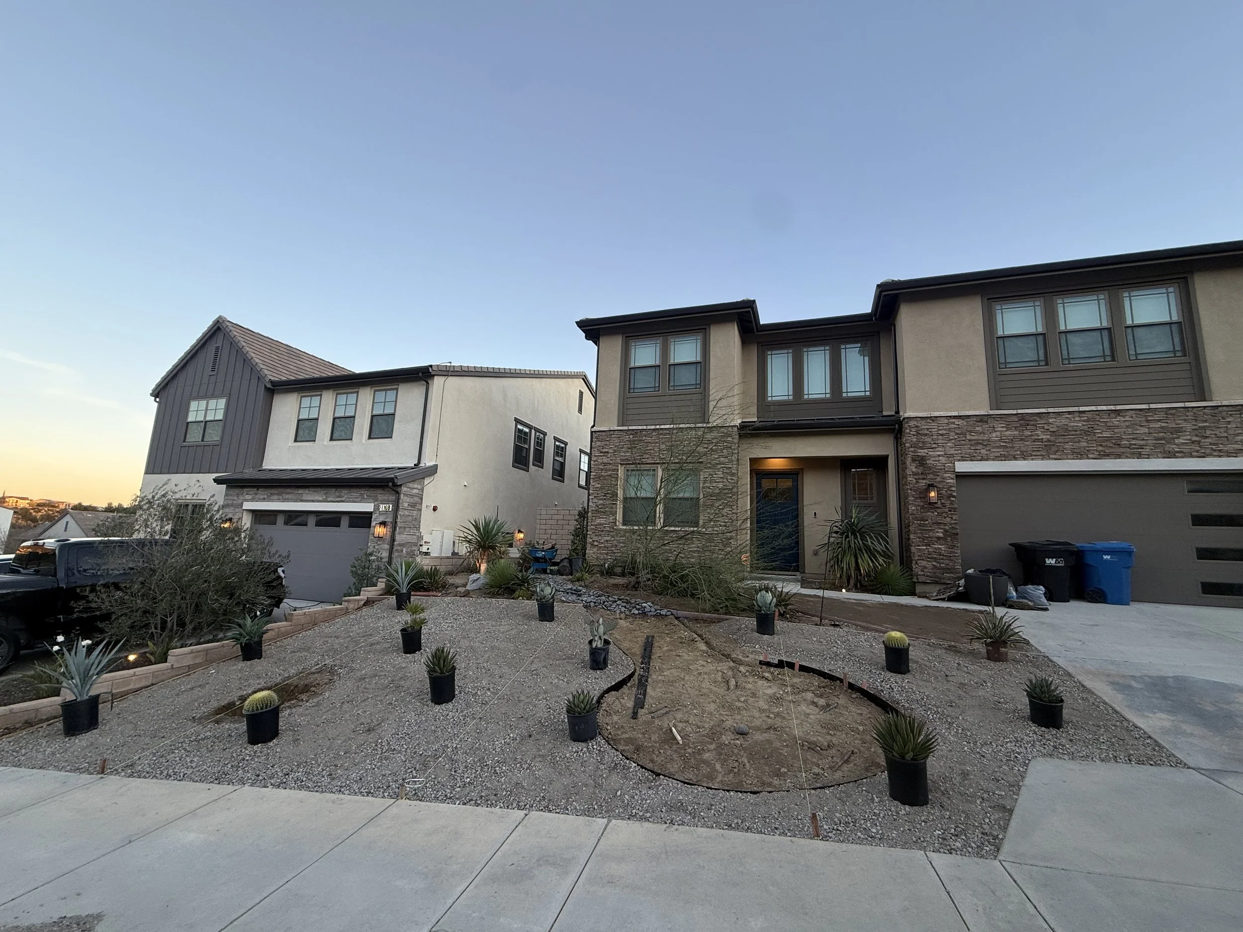Front view of modern multi-story residential buildings with landscaped front yard, some potted plants, and construction materials, under a clear blue sky at dusk.