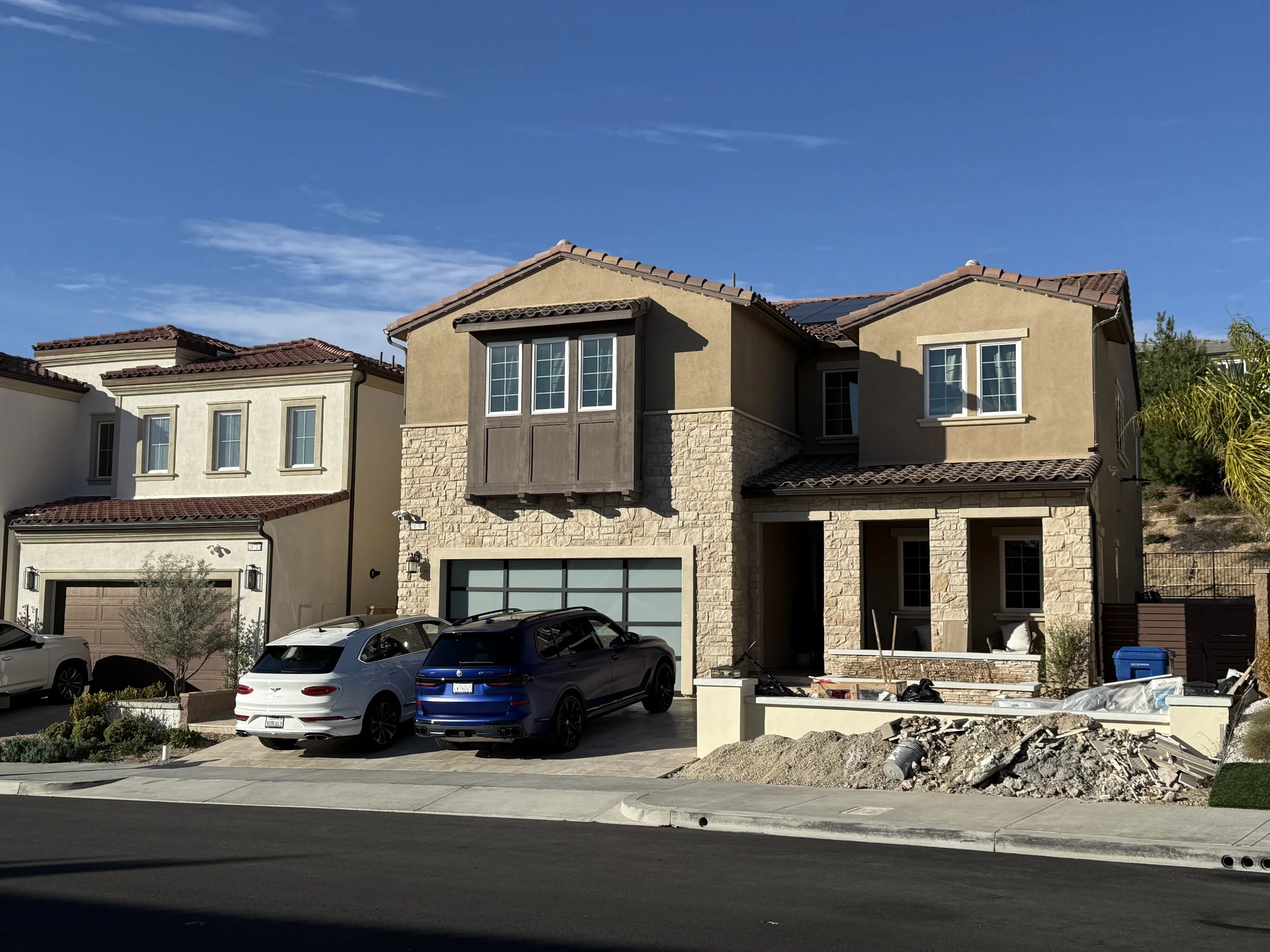 Front view of a row of houses, with cars parked in the driveway, under a blue sky. The house in the center is under construction, with unfinished siding and construction debris nearby.