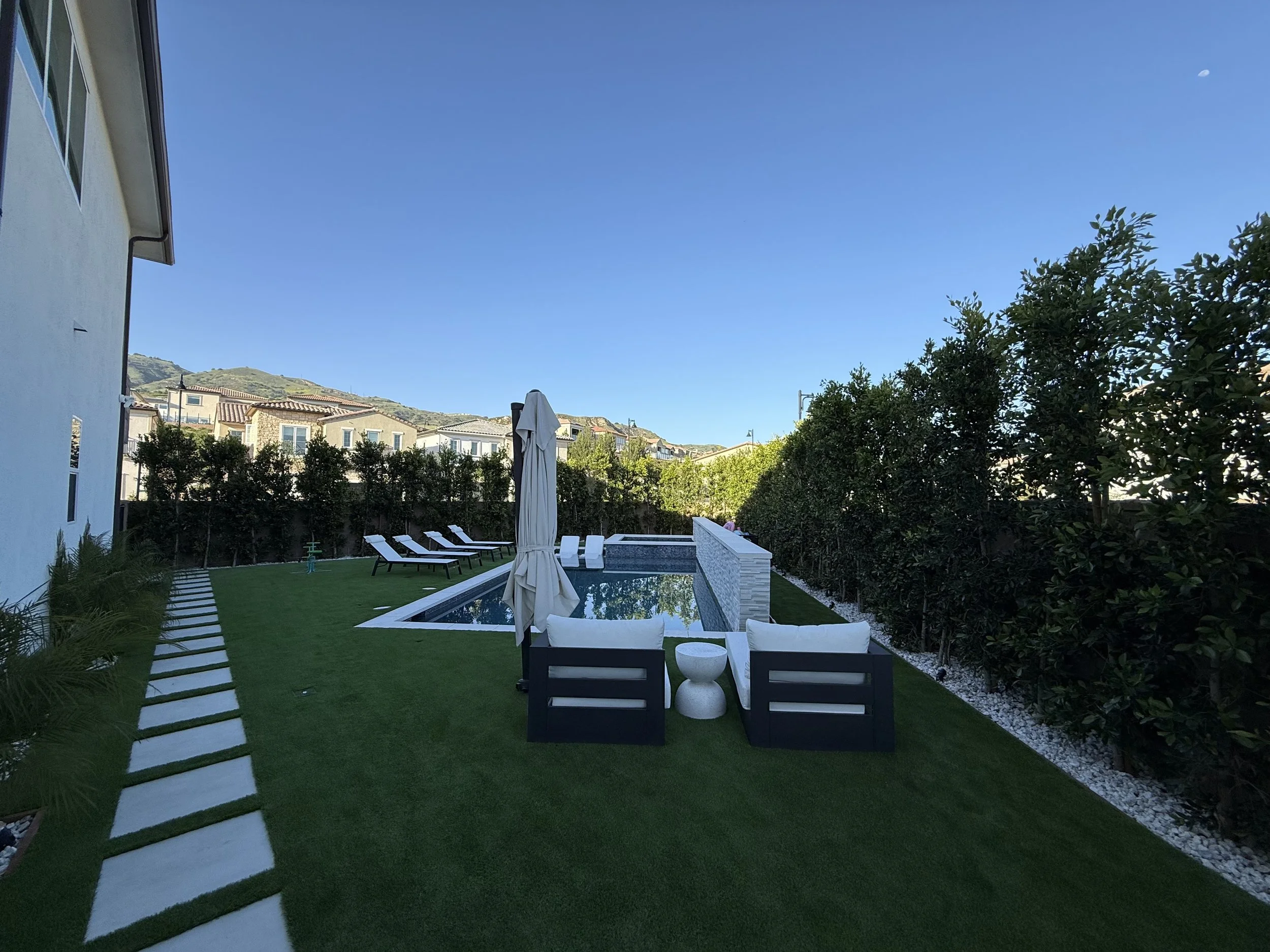 A backyard with a pool, outdoor furniture, an umbrella, and a well-maintained lawn, surrounded by bushes and trees under a clear blue sky.