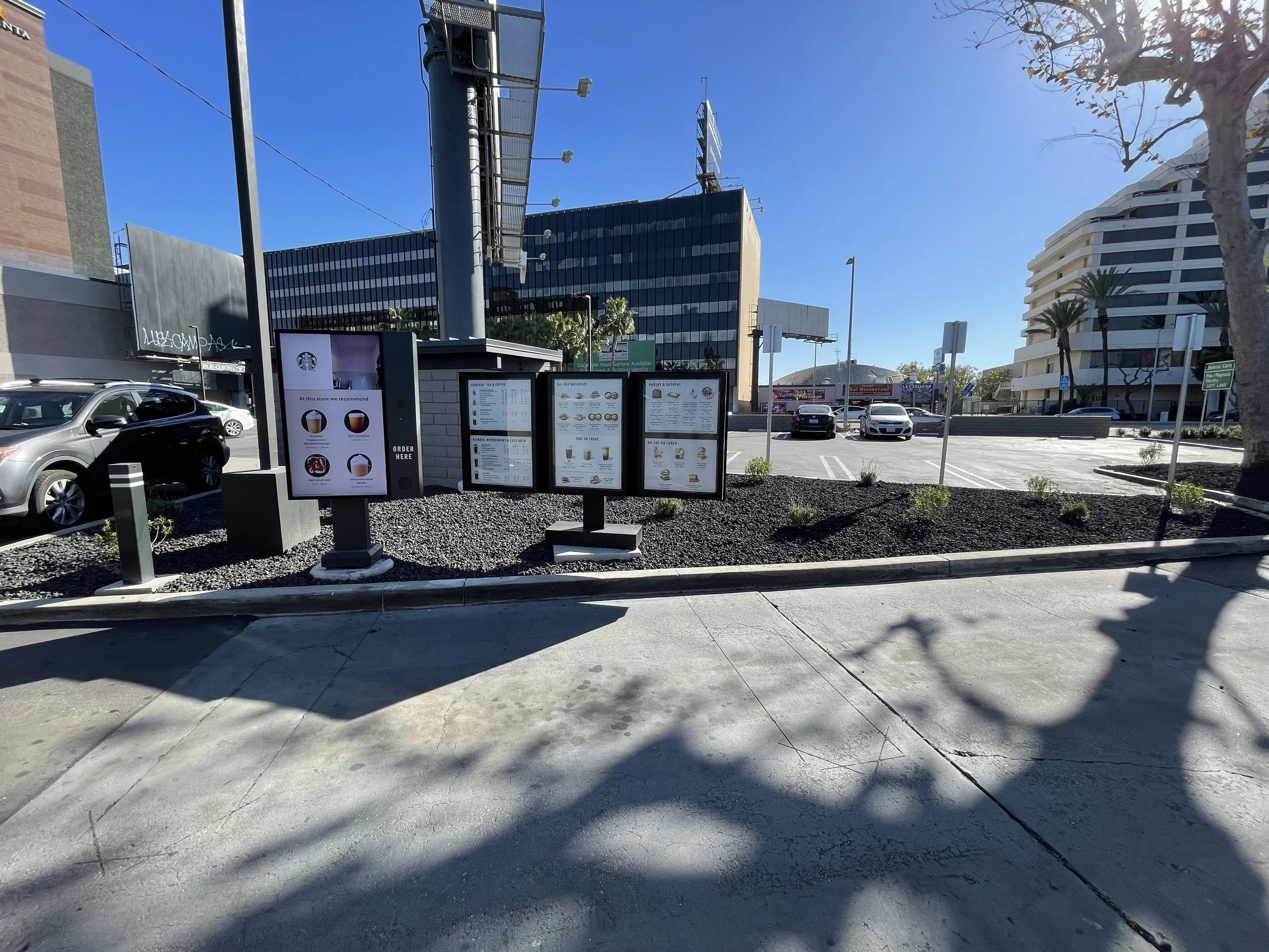 A clear blue sky atop a city scene with a parking lot, modern office buildings, and billboards. There are trees, parked cars, and outdoor menu boards for a restaurant or cafe.