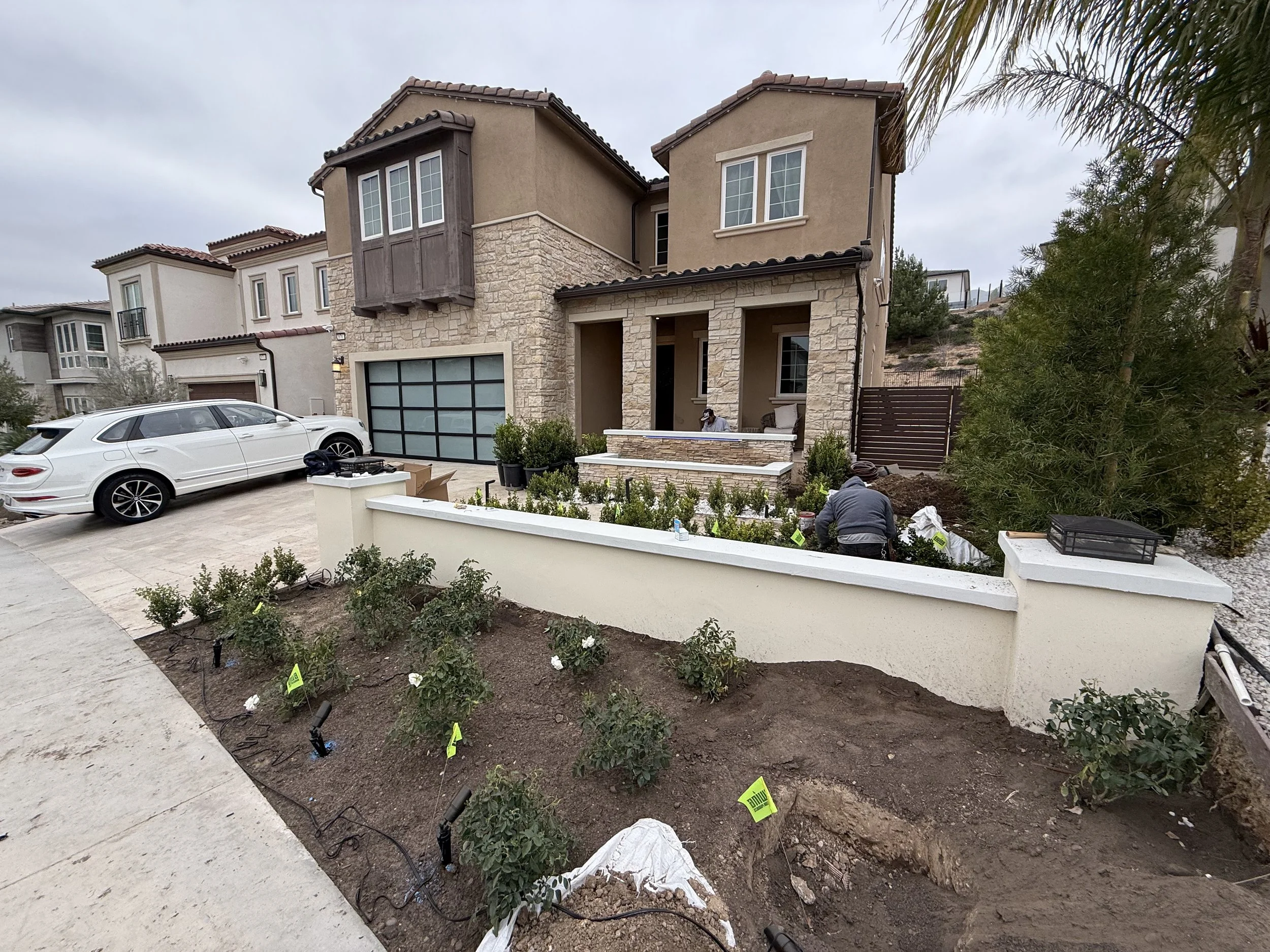 A house under construction with a front yard where plants are being planted, workers, and construction equipment visible. The house has a modern style with stone and stucco exterior and a driveway with a white car parked.