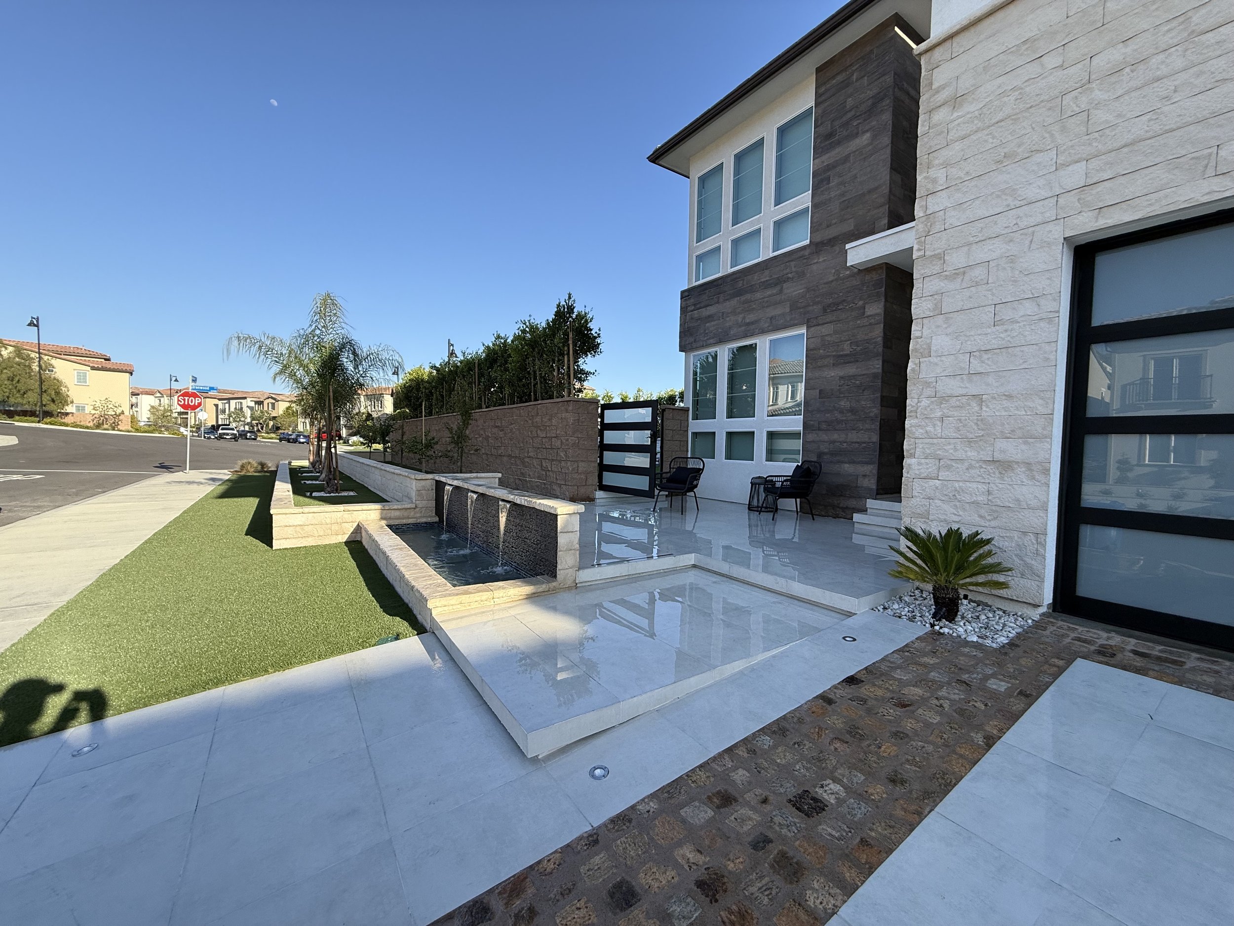 Modern house exterior with white and stone walls, large windows, outdoor seating, and a landscaped yard with a water feature and trees