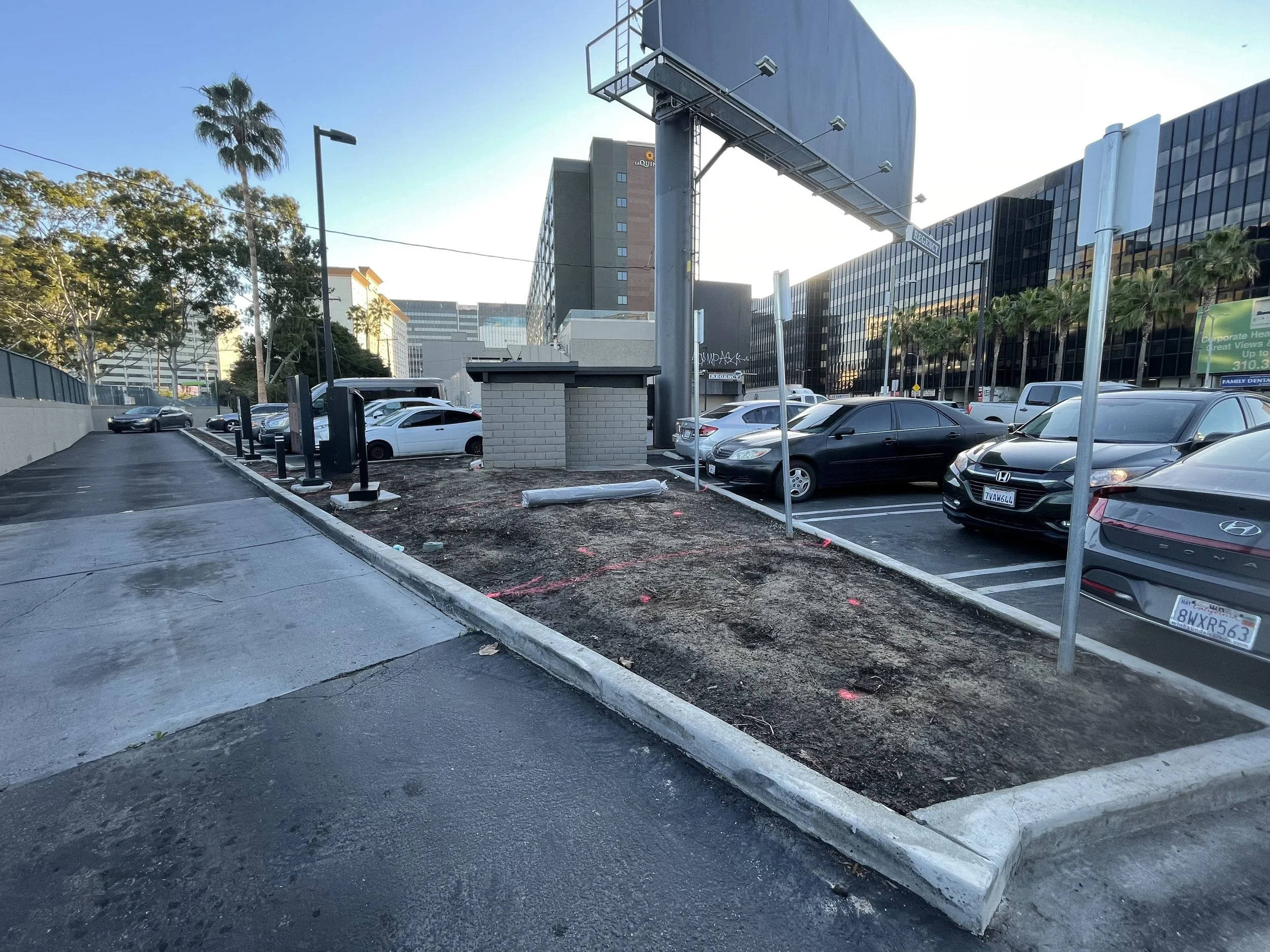 A parking lot with several parked cars, some construction work or repairs ongoing in a small dirt area, a large building with glass windows, and a blue sky with a few clouds.