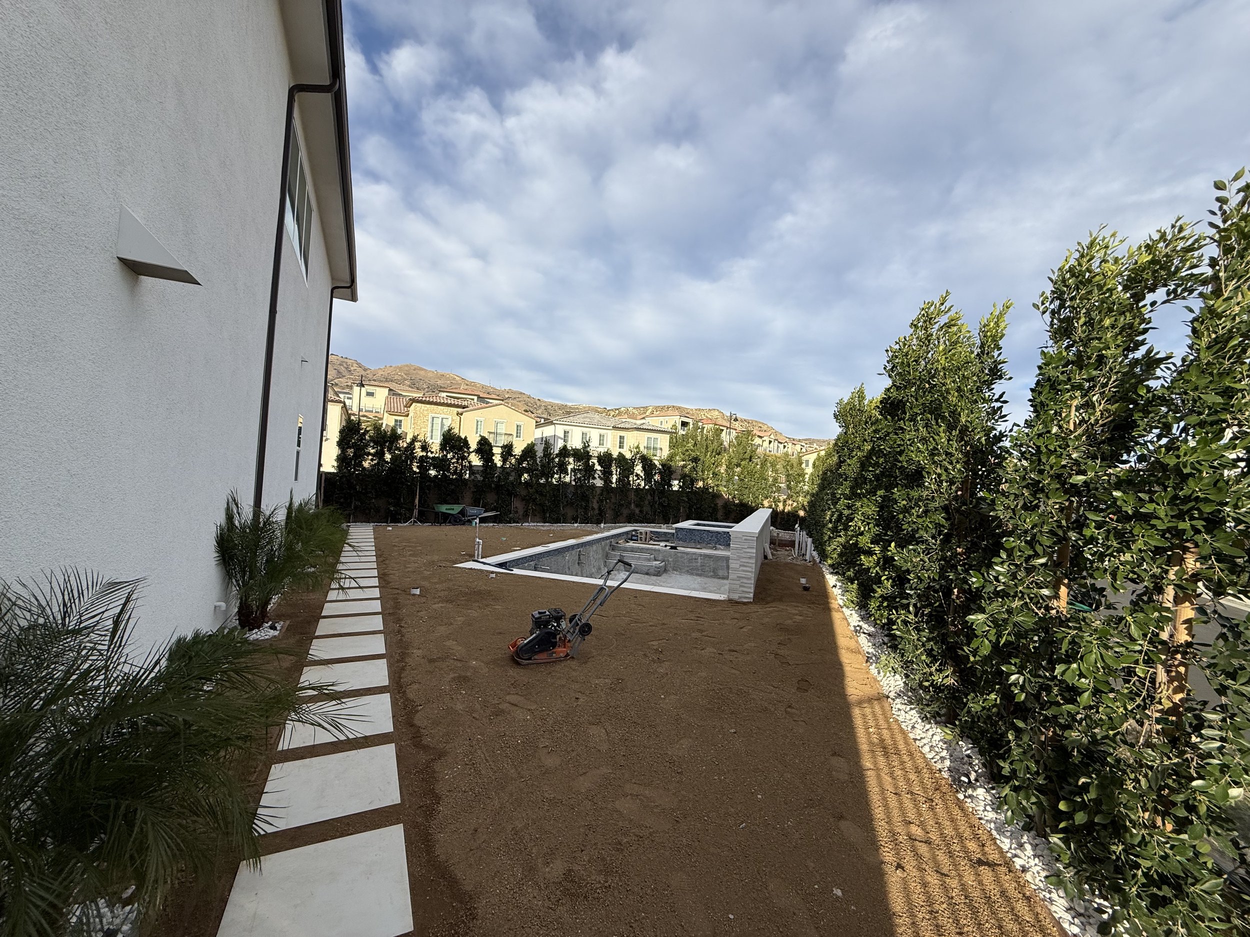 A backyard with a brand new pool installation site, a small hand concrete mixer, and a path of concrete stepping stones along the white house wall, with trees and neighboring houses in the background.