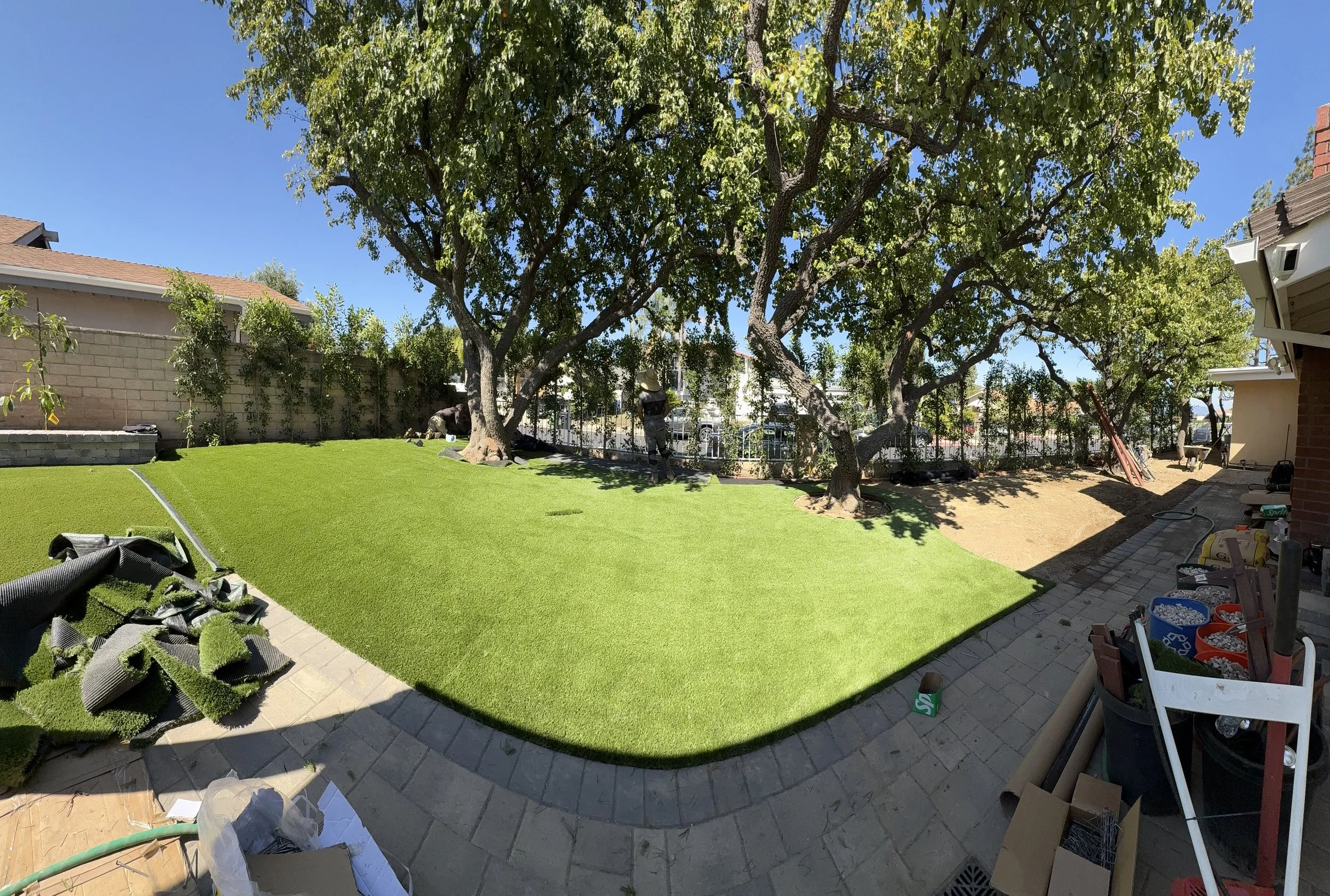 A backyard with a large tree providing shade, a patch of new artificial turf, and construction supplies and tools scattered around as someone works on landscaping or yard renovation.