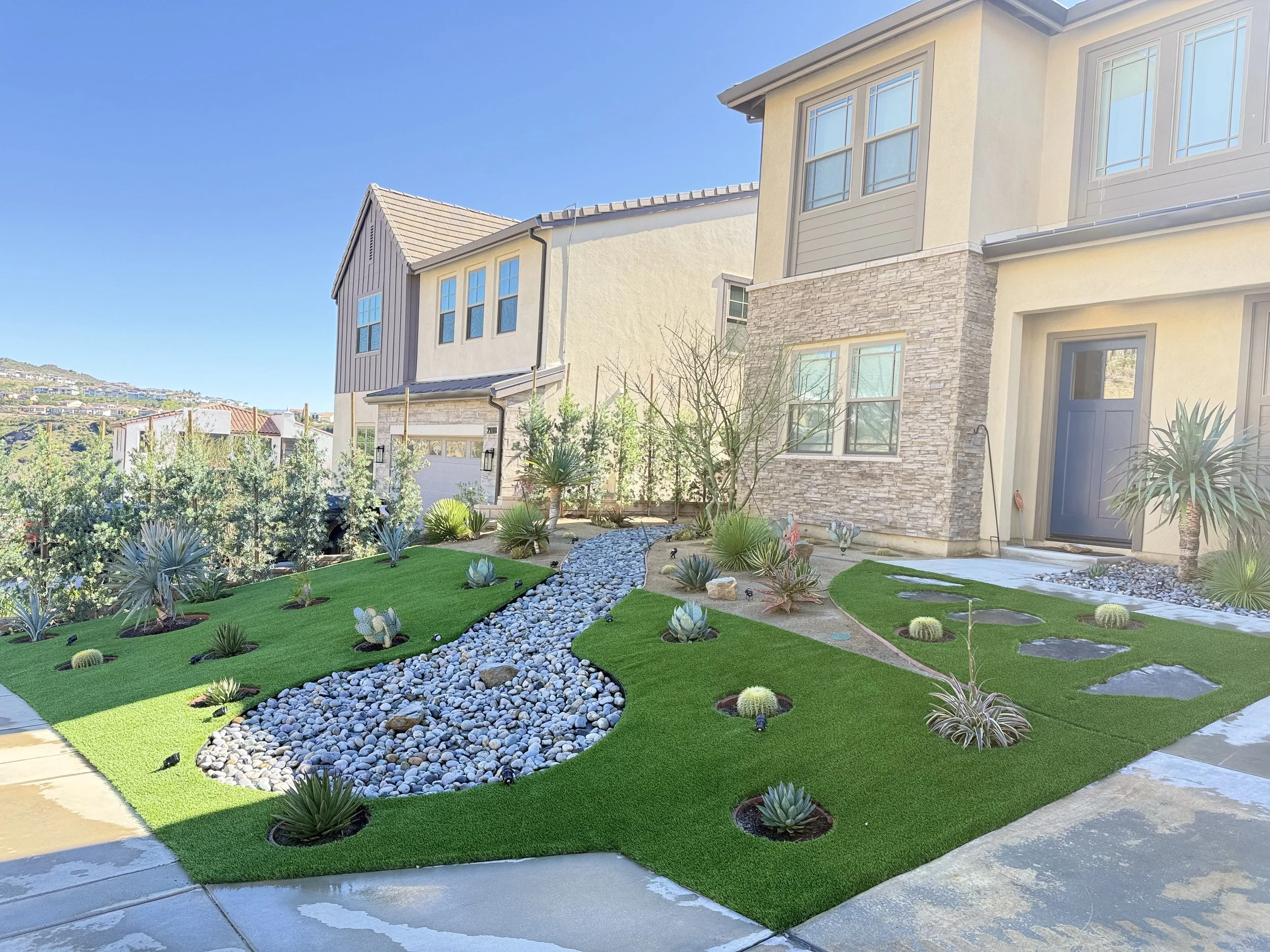 Modern suburban house with a meticulously landscaped front yard featuring artificial grass, decorative rocks, and desert plants under a clear blue sky.