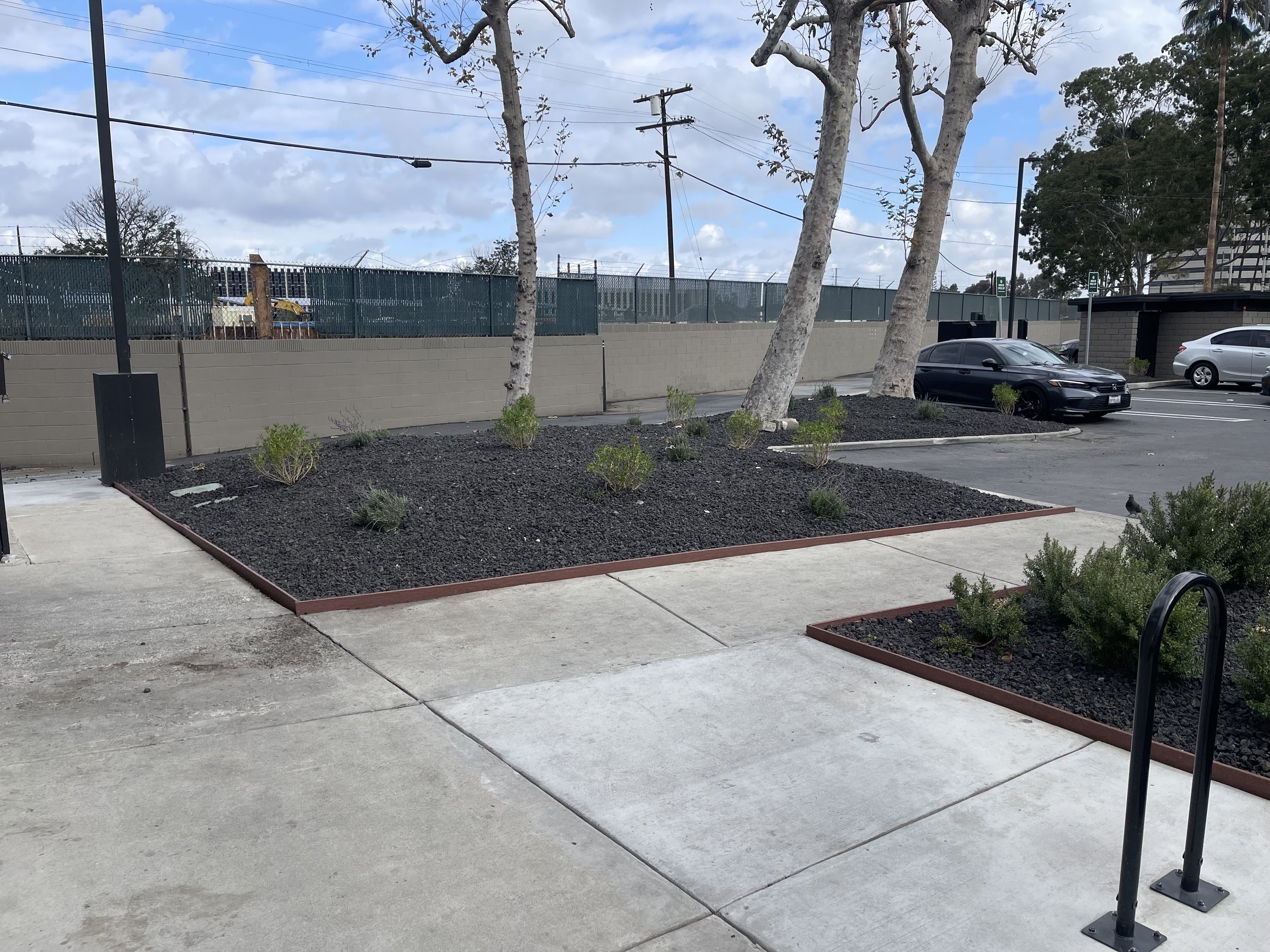 View of a parking lot with a few parked cars, trees, a concrete wall topped with a green fence, utility poles, and a partly cloudy sky.