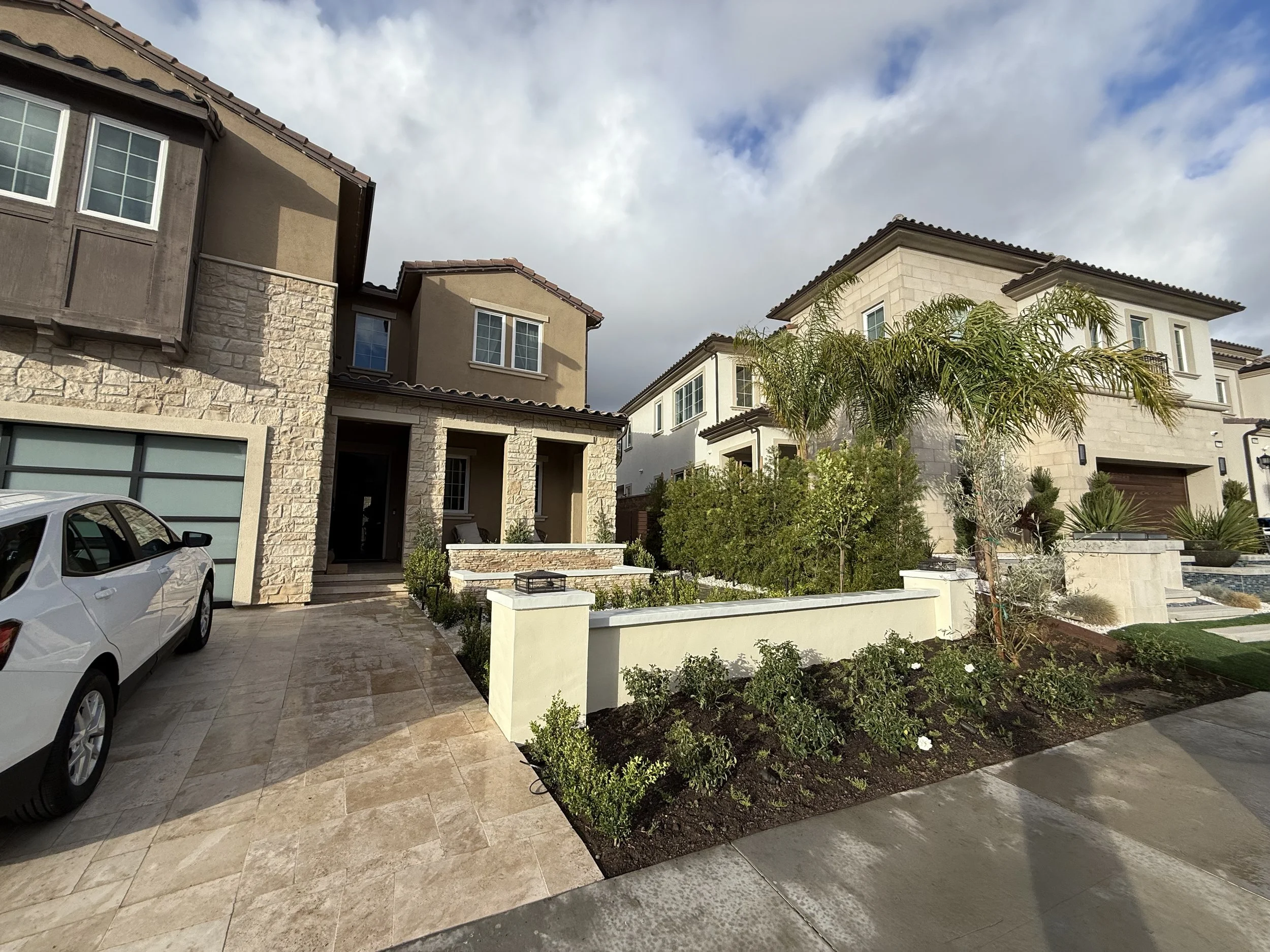 Front yard view of modern two-story houses with tiled roofs, large windows, and landscaped lawns with small trees and shrubs. A white car is parked in the driveway, and the sky is partly cloudy.