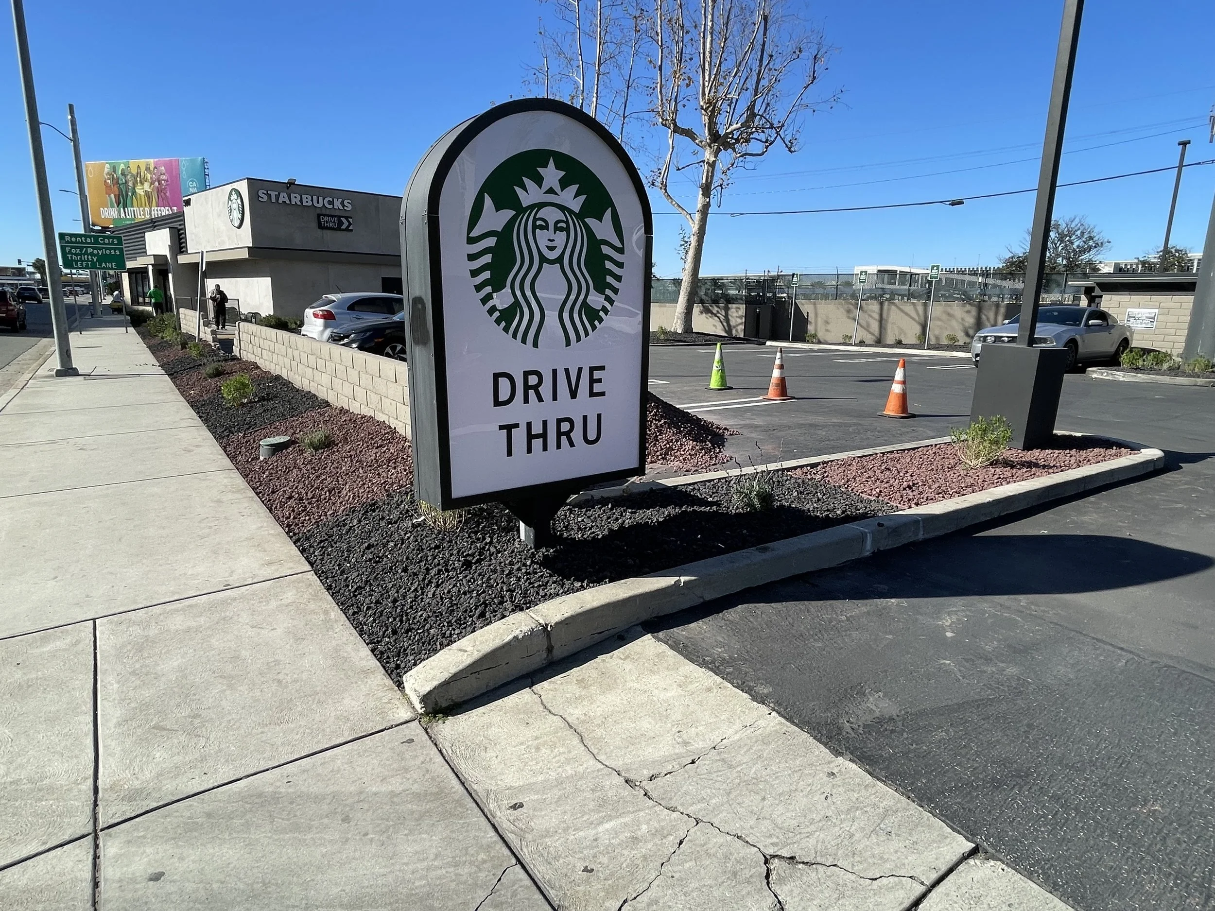 A Starbucks drive-thru sign in a parking lot with parked cars and a clear blue sky.