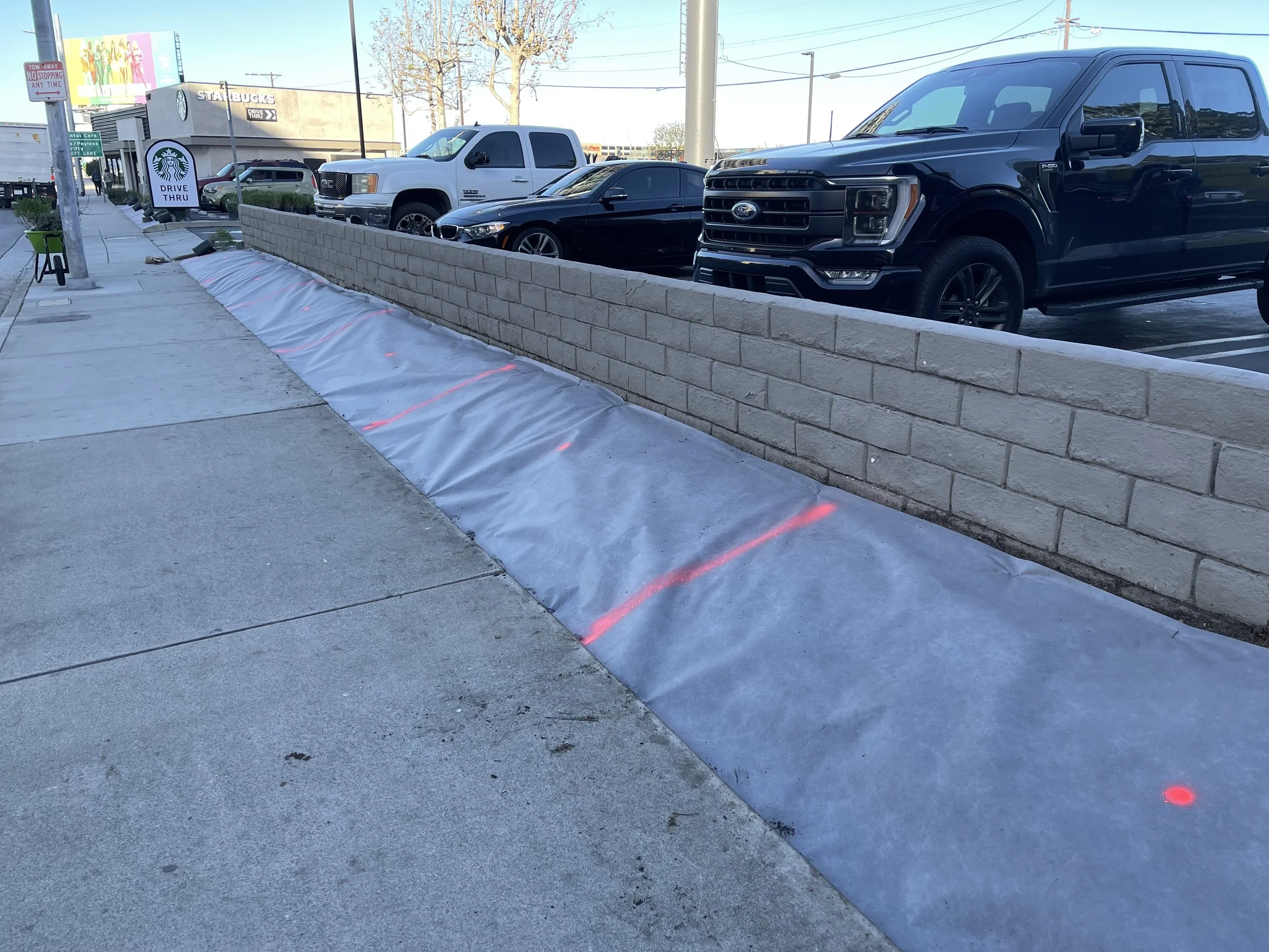 A sidewalk with a low brick wall separating it from a parking lot with several cars. There is a sign indicating a Starbucks drive-thru in the background and a parking lot with cars in front of a retail store. A small green trash bin is near the sidew
