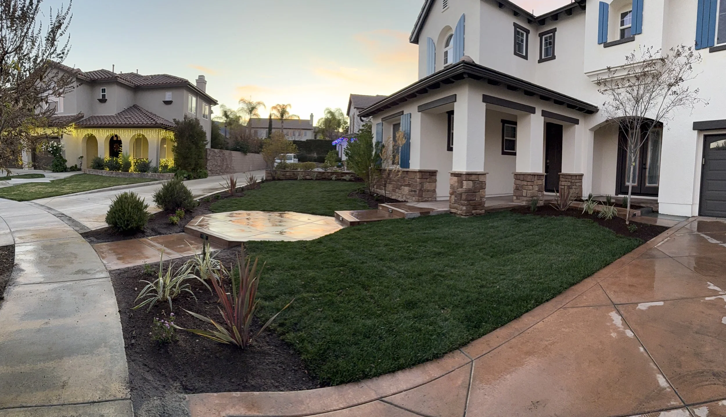Residential backyard with manicured lawn, stone pathway, and white house with blue shutters at sunset.