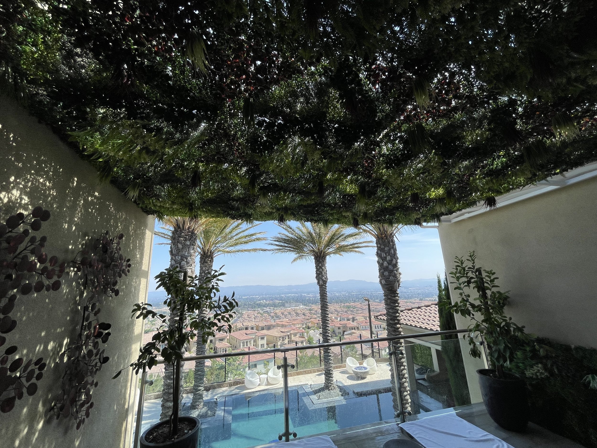 A house or balcony with a swimming pool, surrounded by trees and plants, with a view of a residential neighborhood and mountains in the distance.