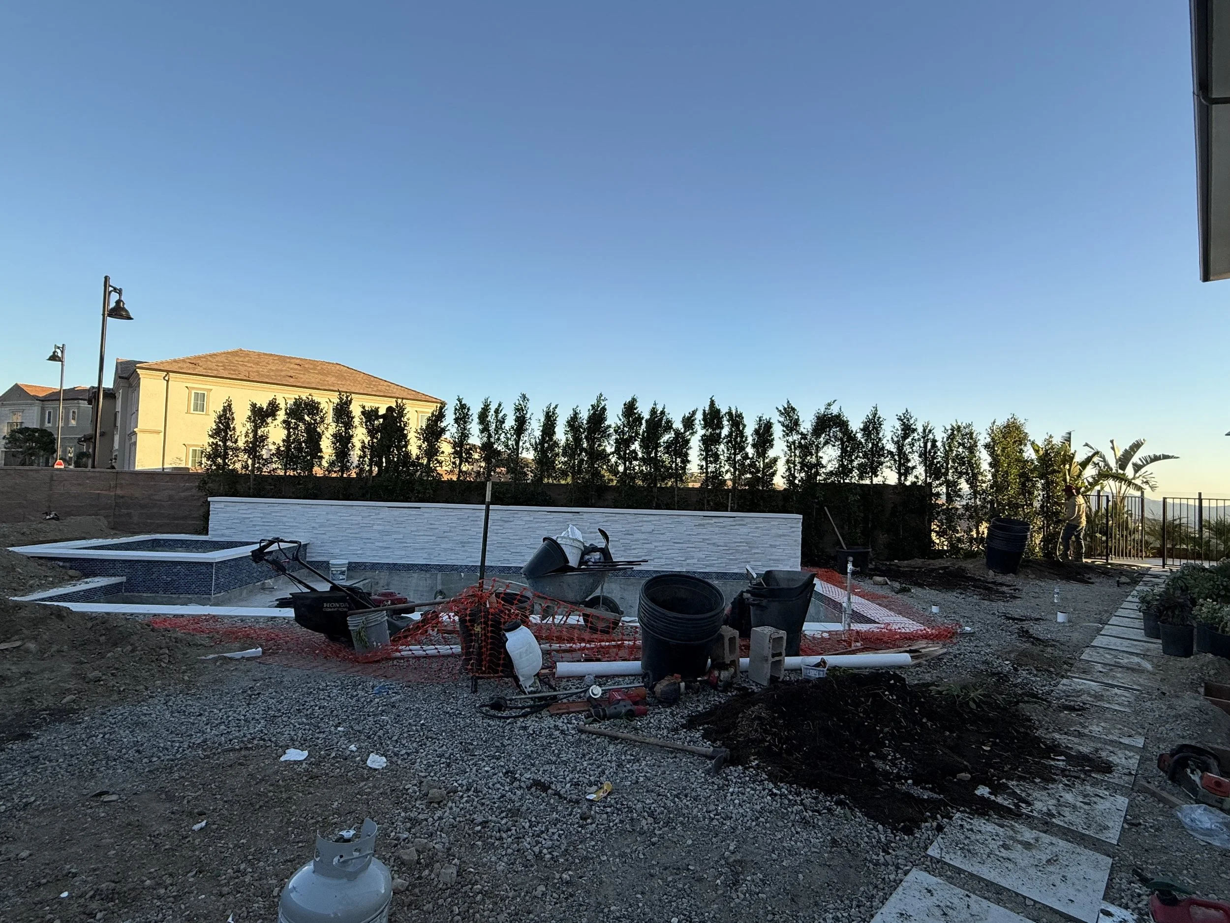 Construction site with gravel ground, construction tools, debris, and a small pool in the foreground. In the background, there are houses, trees, streetlights, and a clear blue sky.